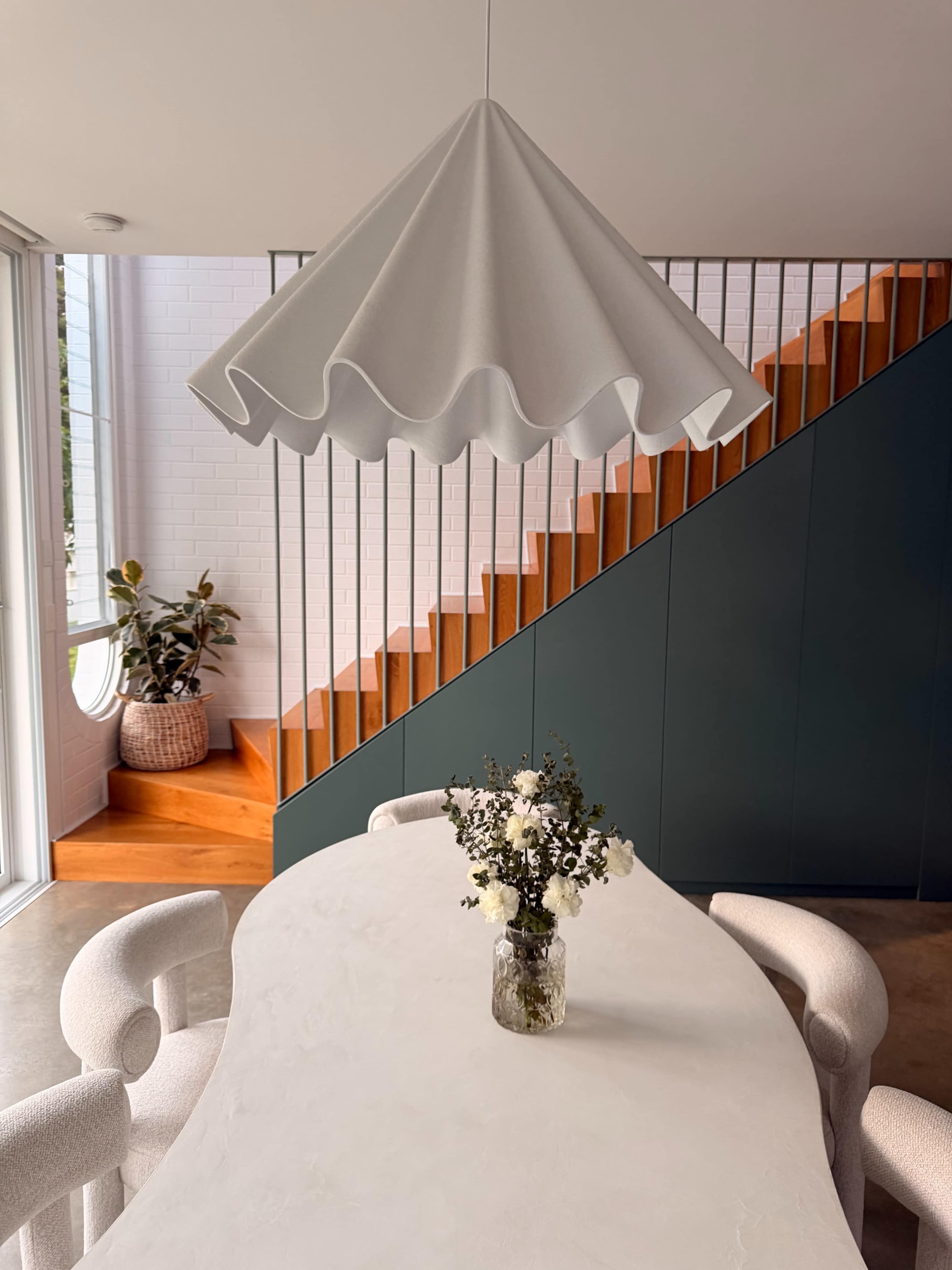 A white, scalloped pendant light hangs above a round table adorned with a floral arrangement, while a wooden staircase and a potted plant are visible in the background.