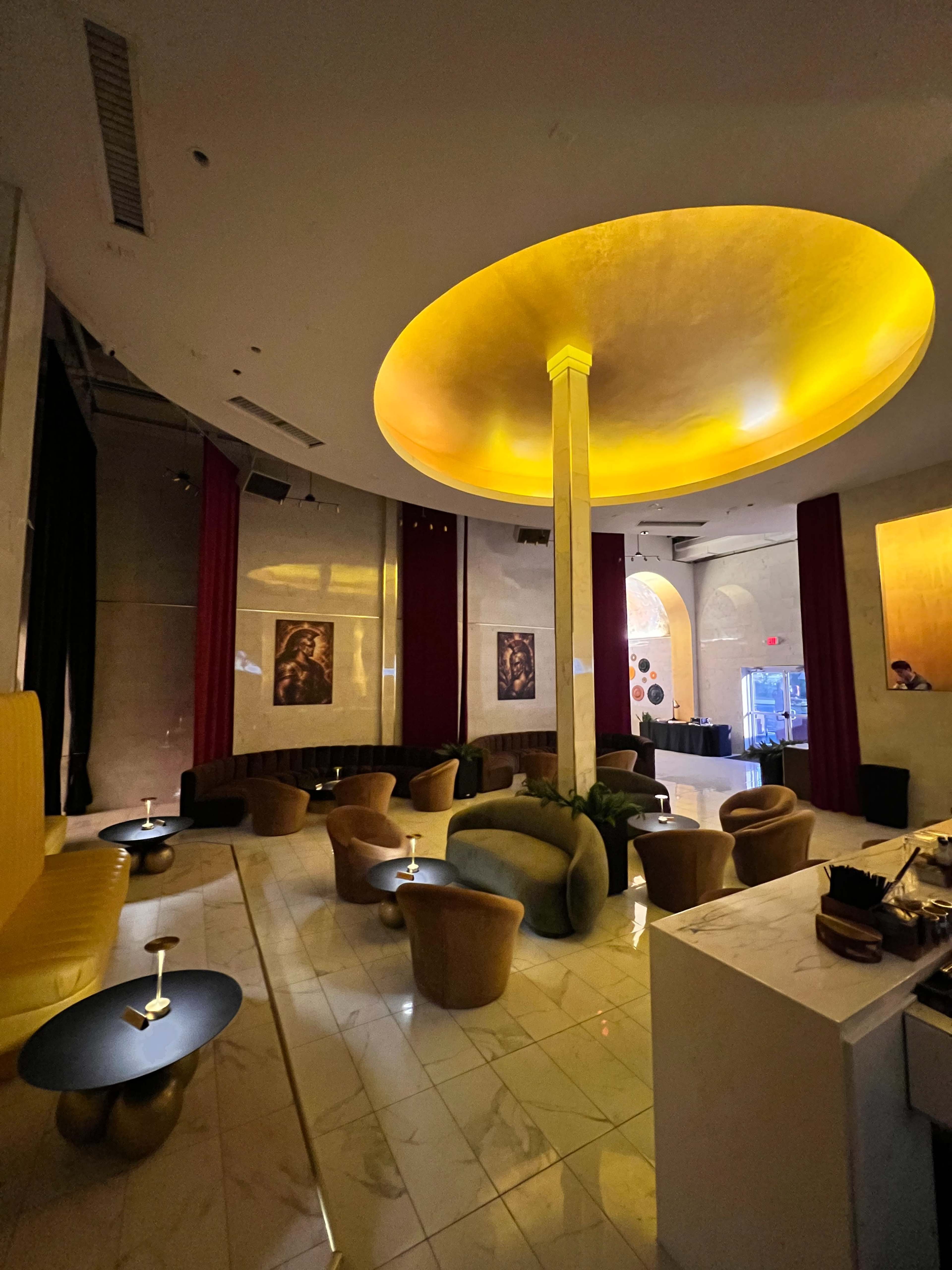 The image shows a modern lounge interior with a circular ceiling featuring warm lighting, plush seating, and a bar area.