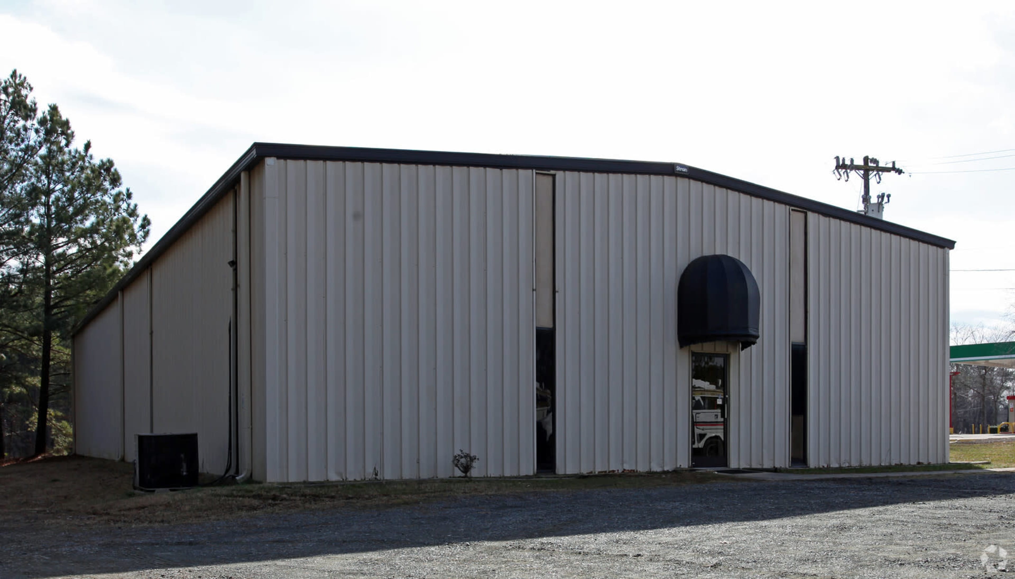 A large, gray metal building with a curved roof is set against a backdrop of trees and power lines.