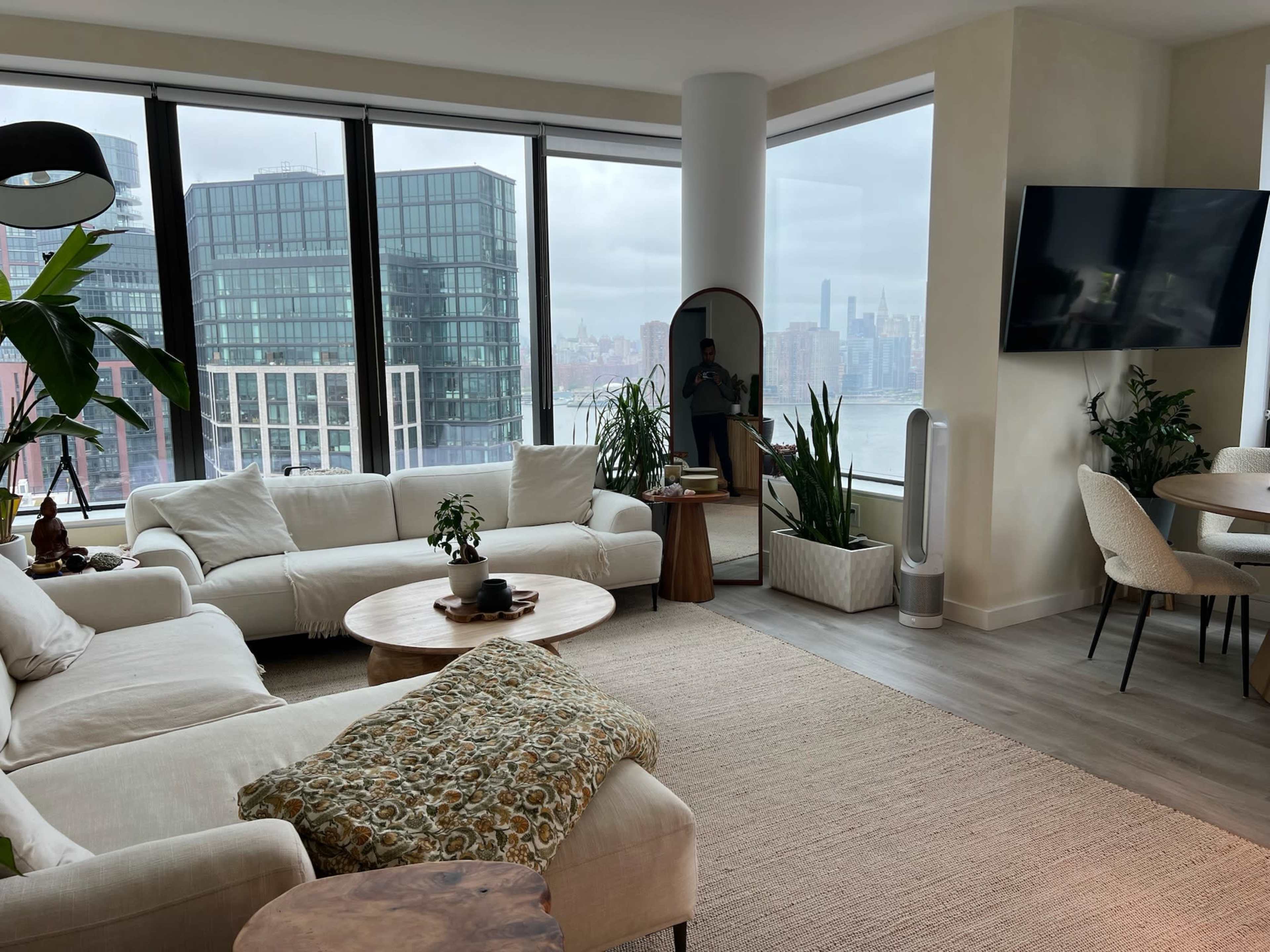 A modern living room features a large sectional sofa, a coffee table, plants, and a view of the city skyline through expansive windows.