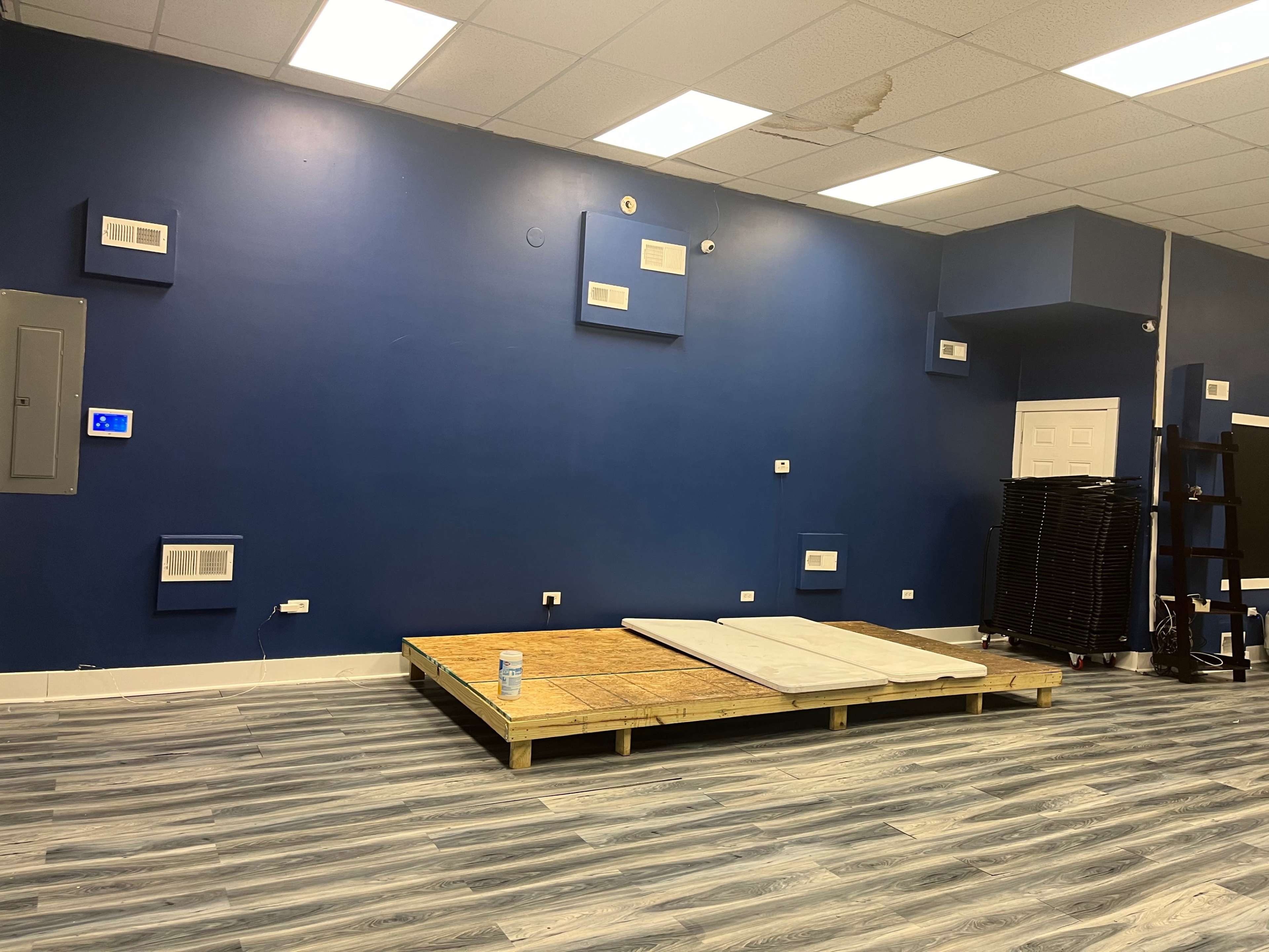 An empty room with a blue wall, a wooden platform with two mattresses on it, and a storage area with black folding chairs along the right side.