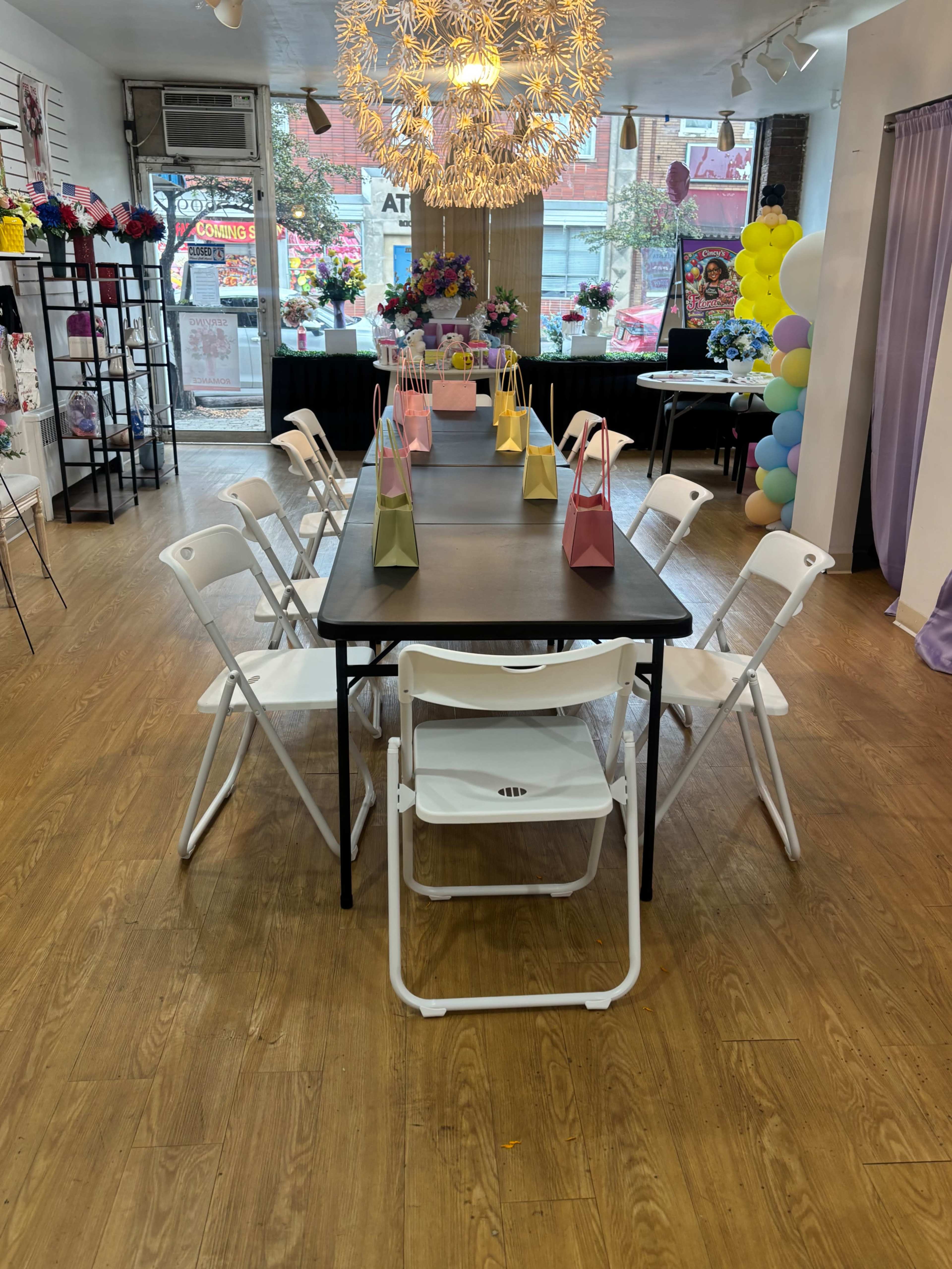 A spacious indoor event area features a long black table surrounded by white folding chairs, decorated with colorful gift bags and a large decorative chandelier.