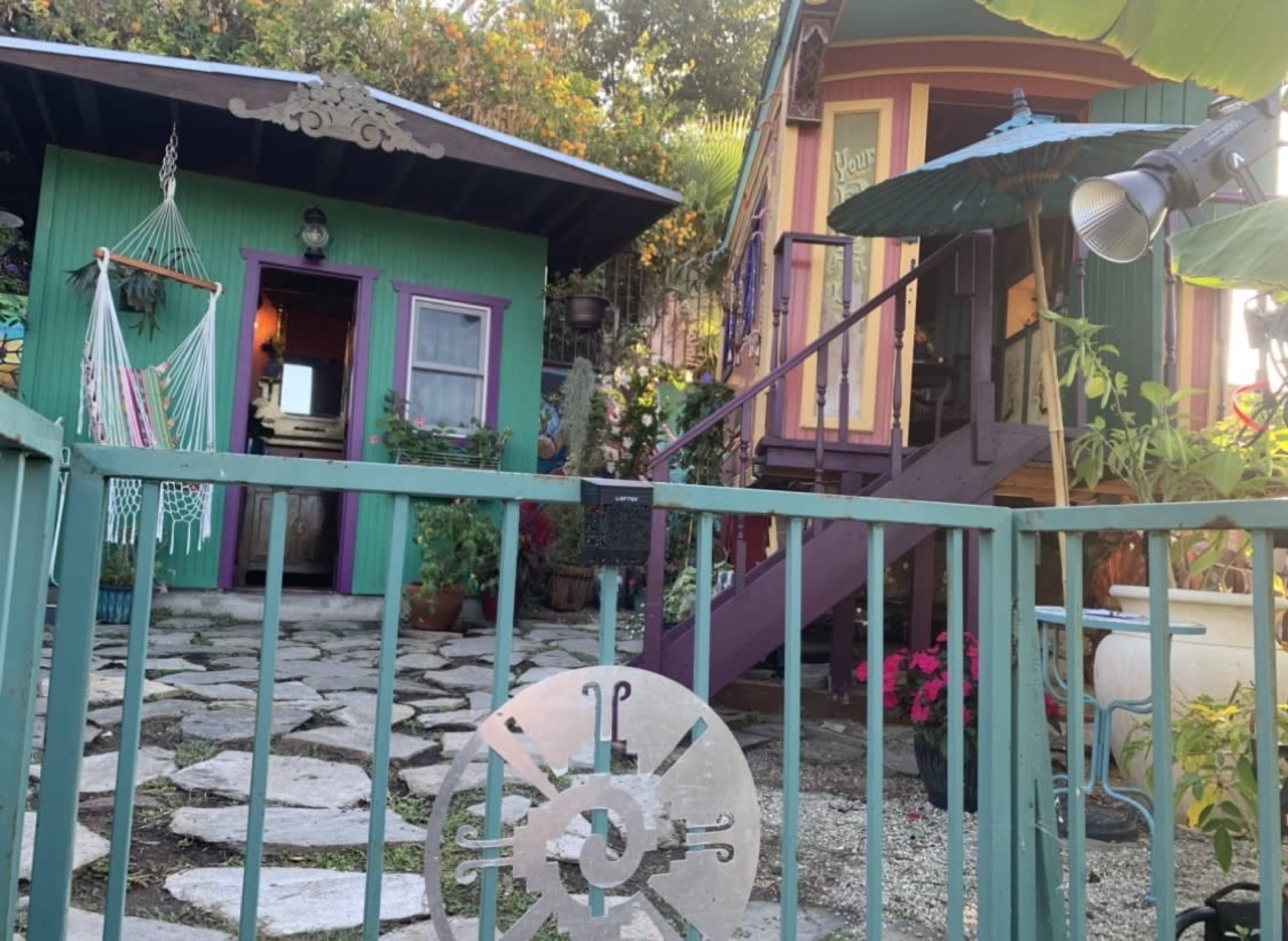 A colorful, small cottage surrounded by lush greenery, with a hammock hanging near a stone pathway leading to the entrance.