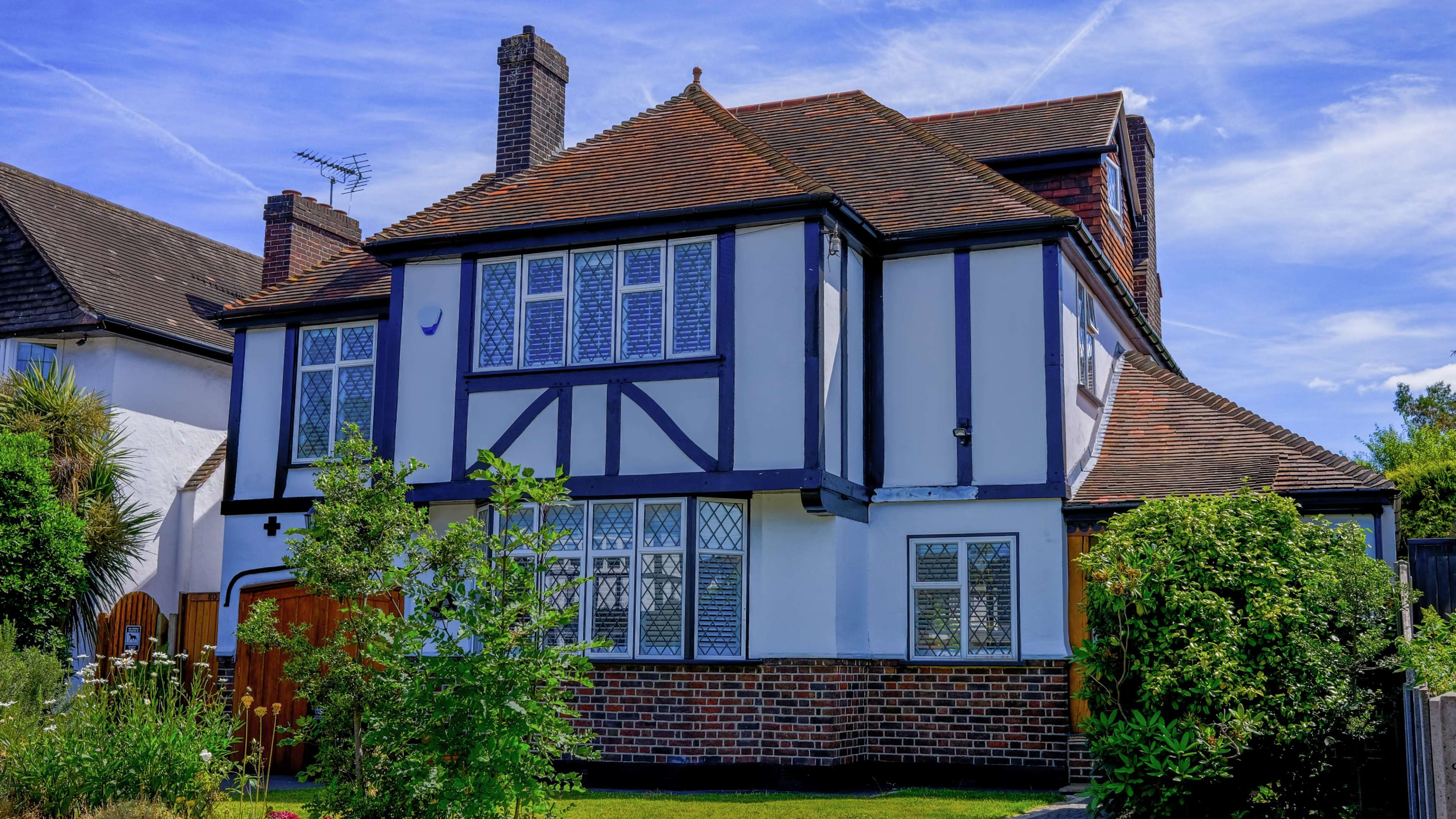 A two-story Tudor-style house with a brick lower level and white upper siding is surrounded by greenery and features multiple leaded-glass windows.