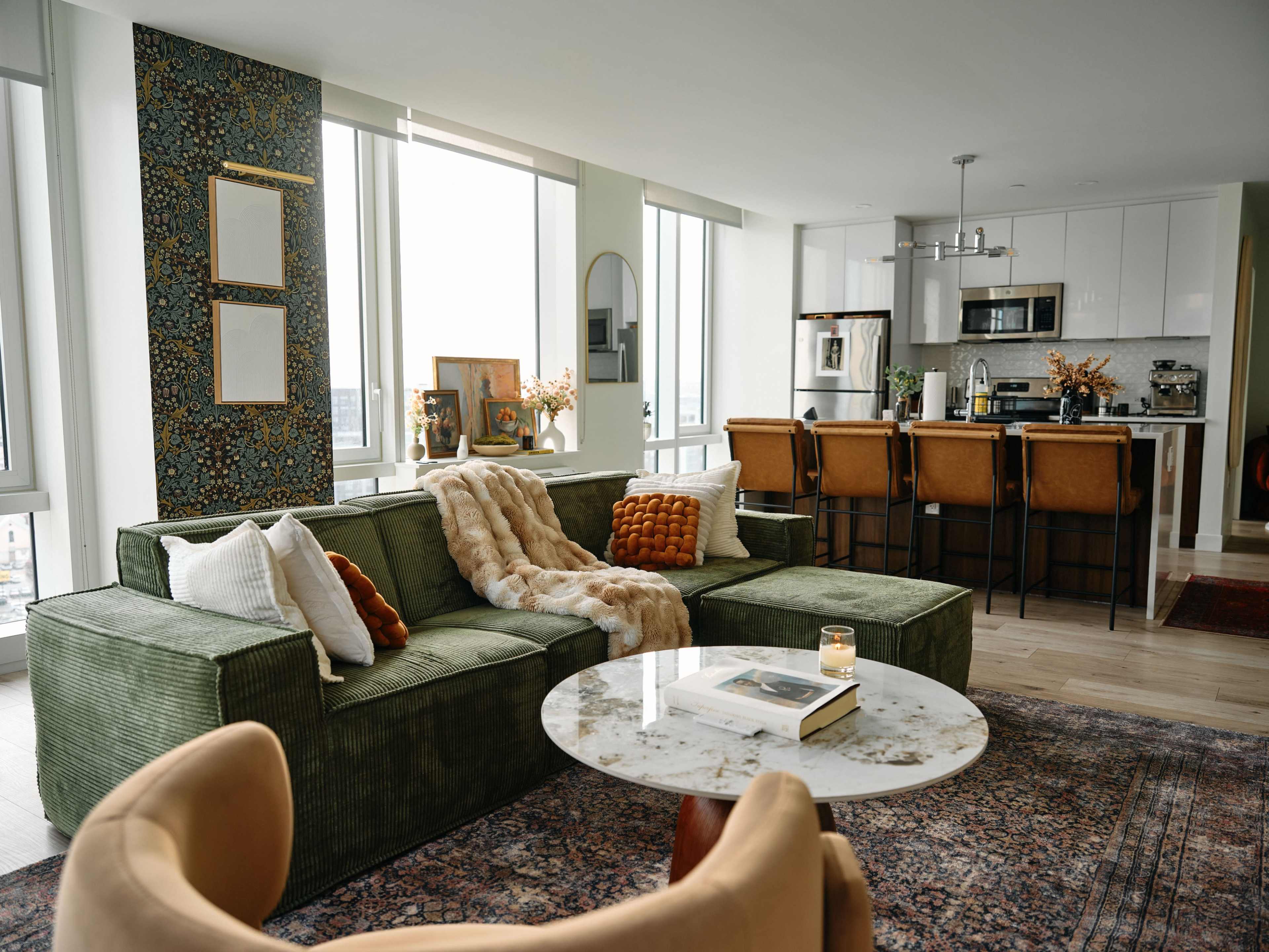 The image shows a modern living room featuring a green sectional sofa, a marble coffee table, and a kitchen area with bar seating in the background.
