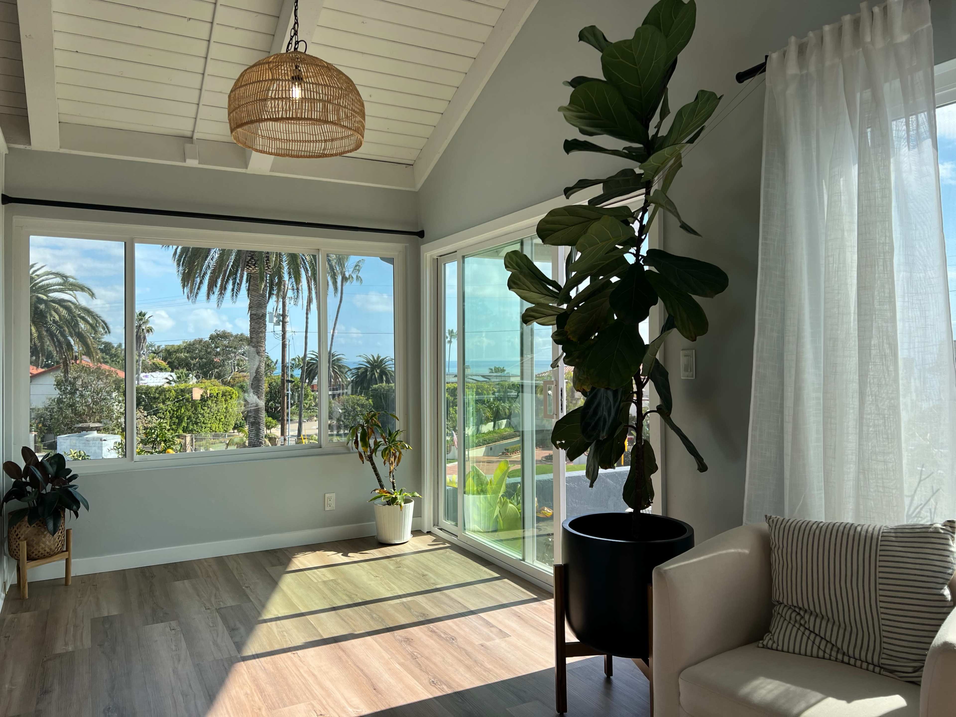 A well-lit room features large windows with a view of palm trees and a waterfront, along with a potted plant and a cozy seating area.