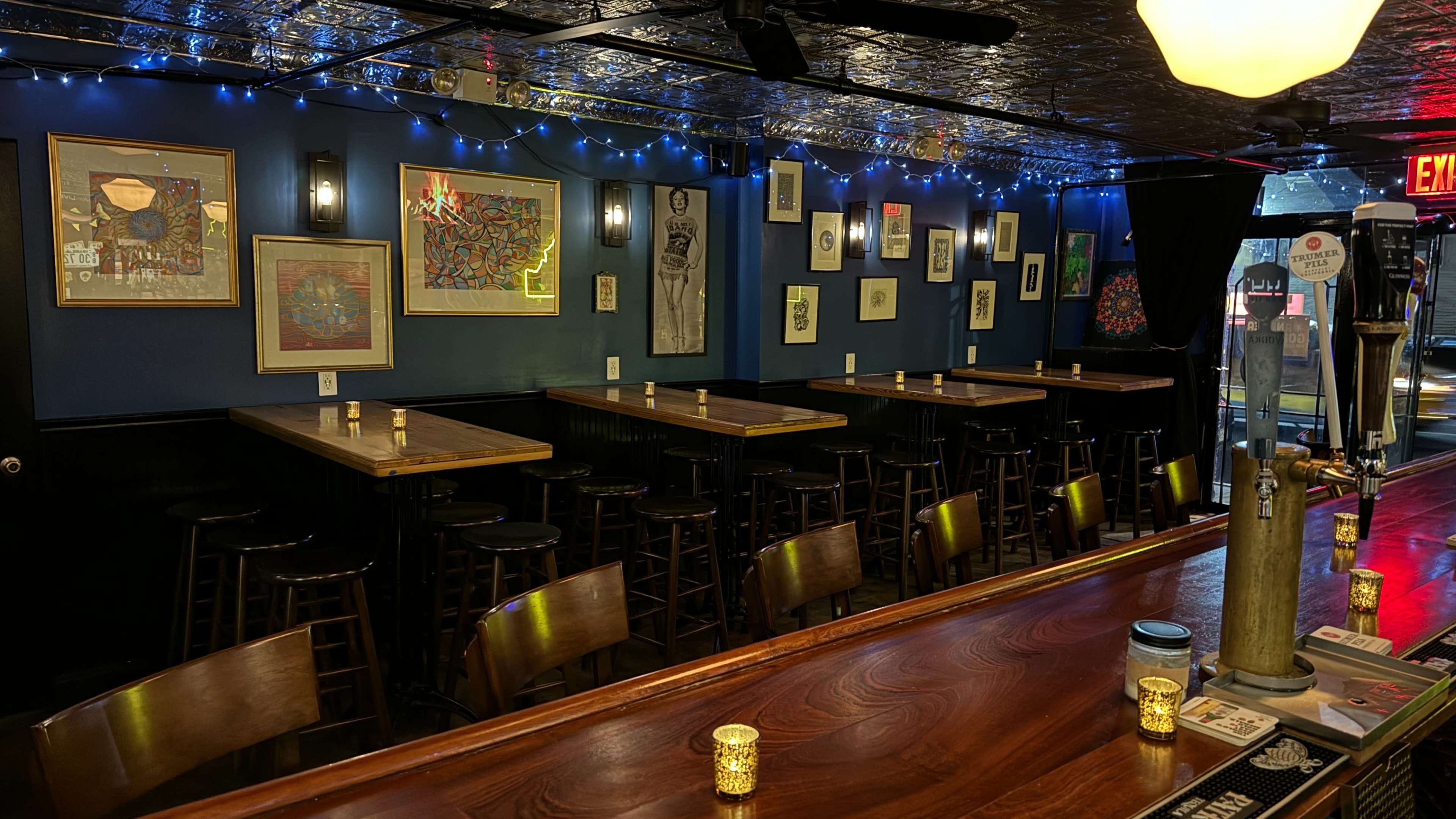 The image shows a dimly lit bar with several tables and stools arranged along the walls, decorated with framed artwork and string lights.