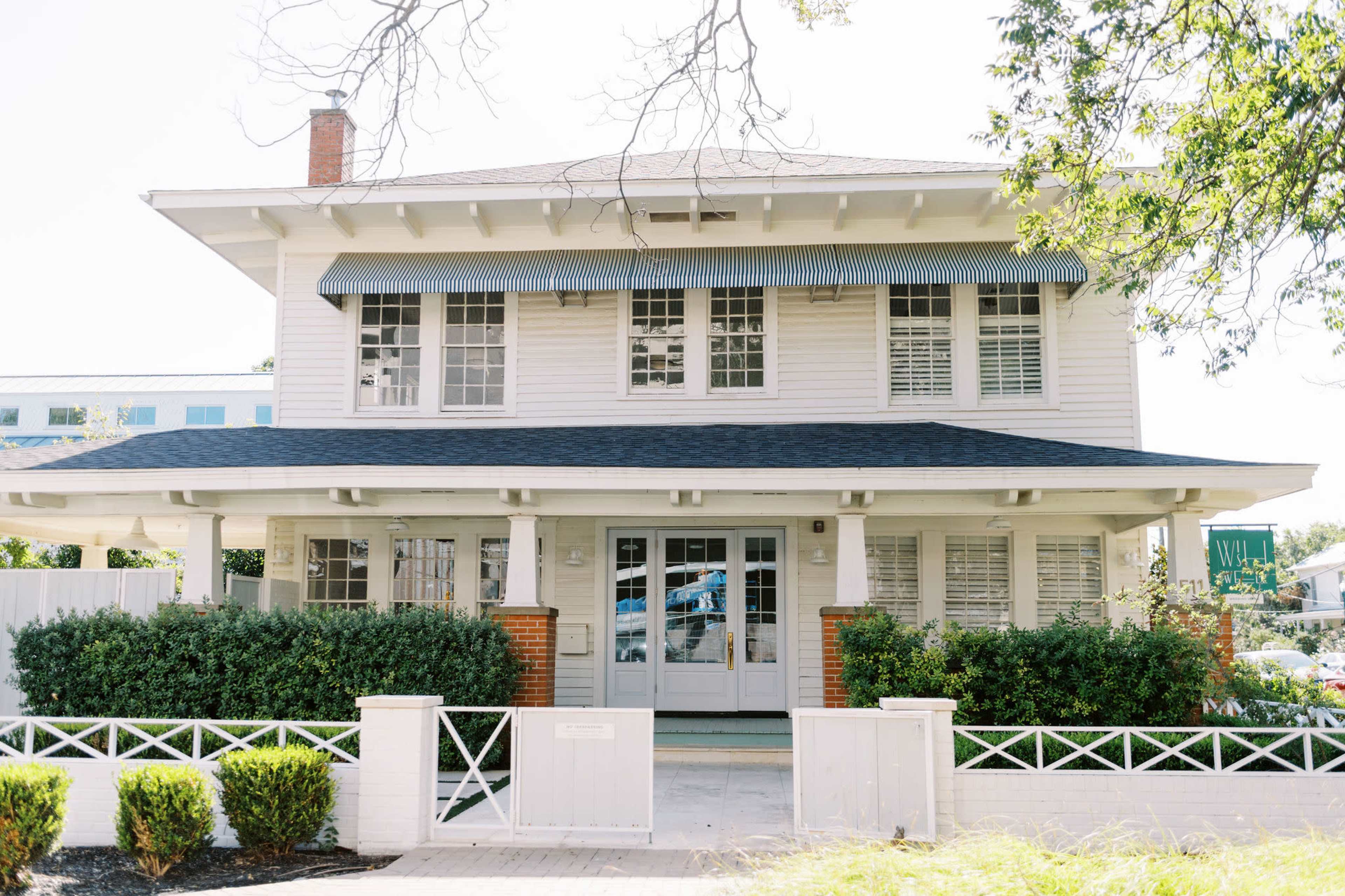 The image shows a two-story white house with a front porch, large windows, and a well-maintained lawn, surrounded by a white fence.