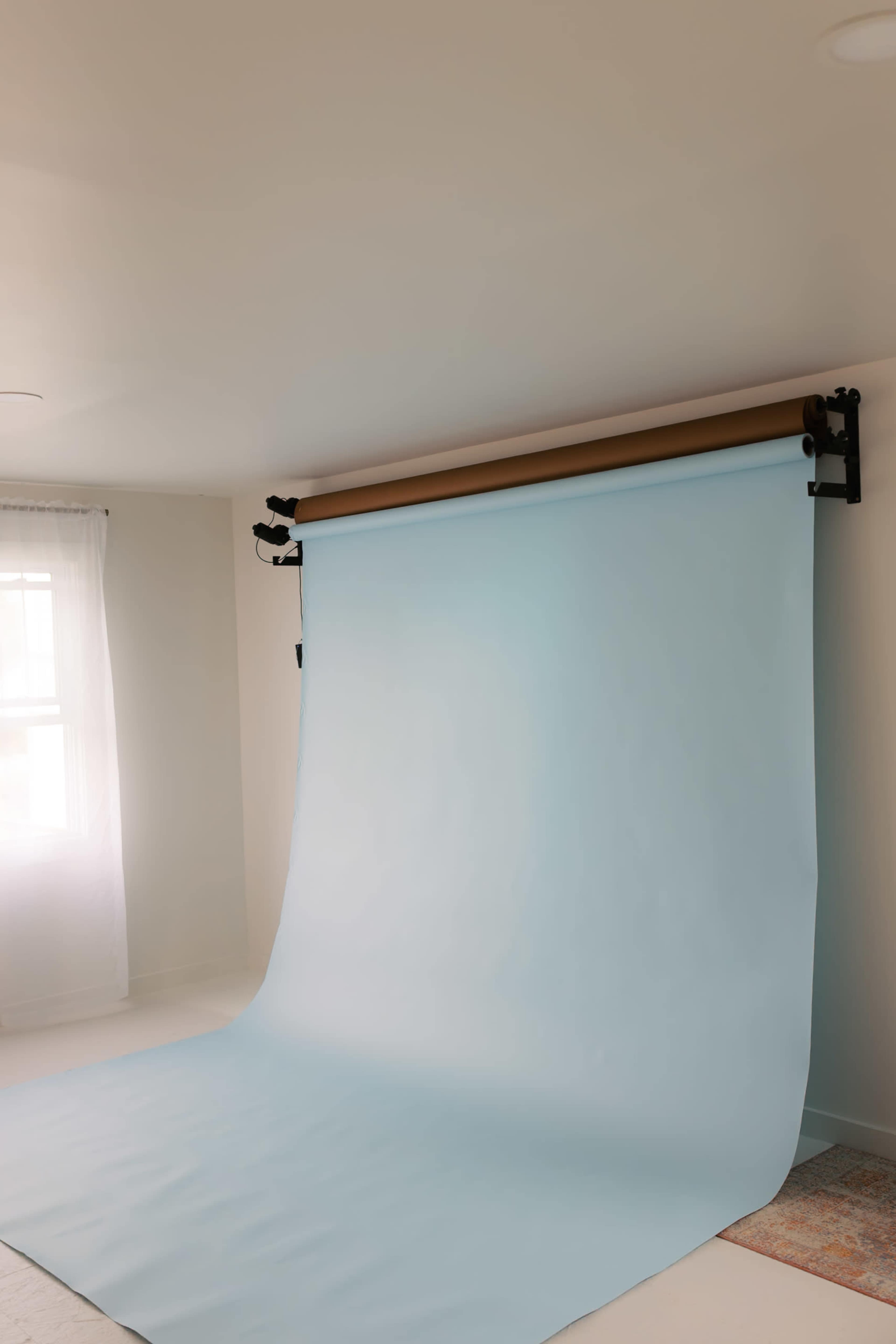 The image shows a light blue backdrop rolled down in a room with neutral-colored walls and a window with sheer curtains.