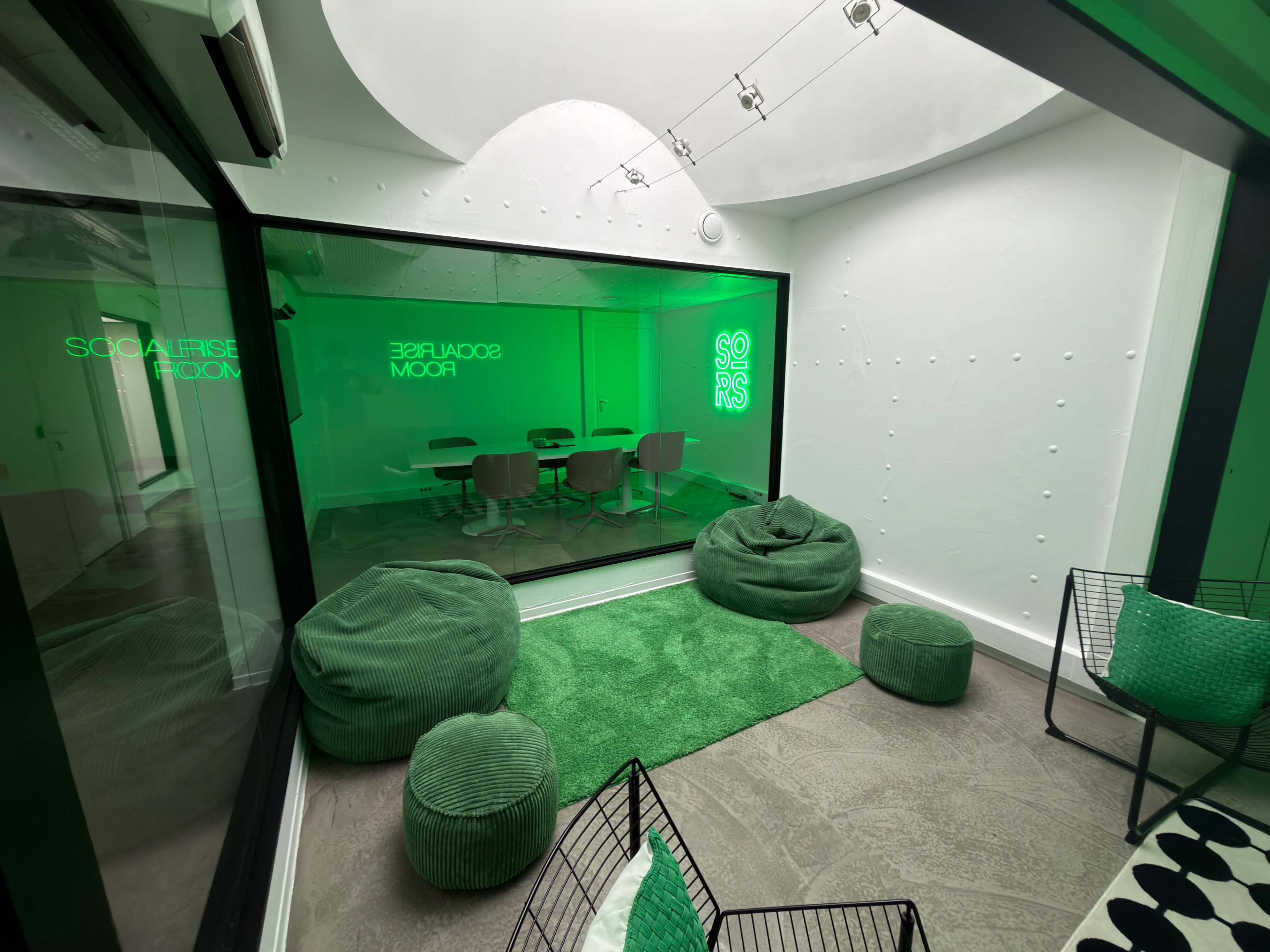The image shows a modern room with green accents, featuring a seating area with bean bags and wire chairs, and a glass-walled conference area containing a table and chairs.