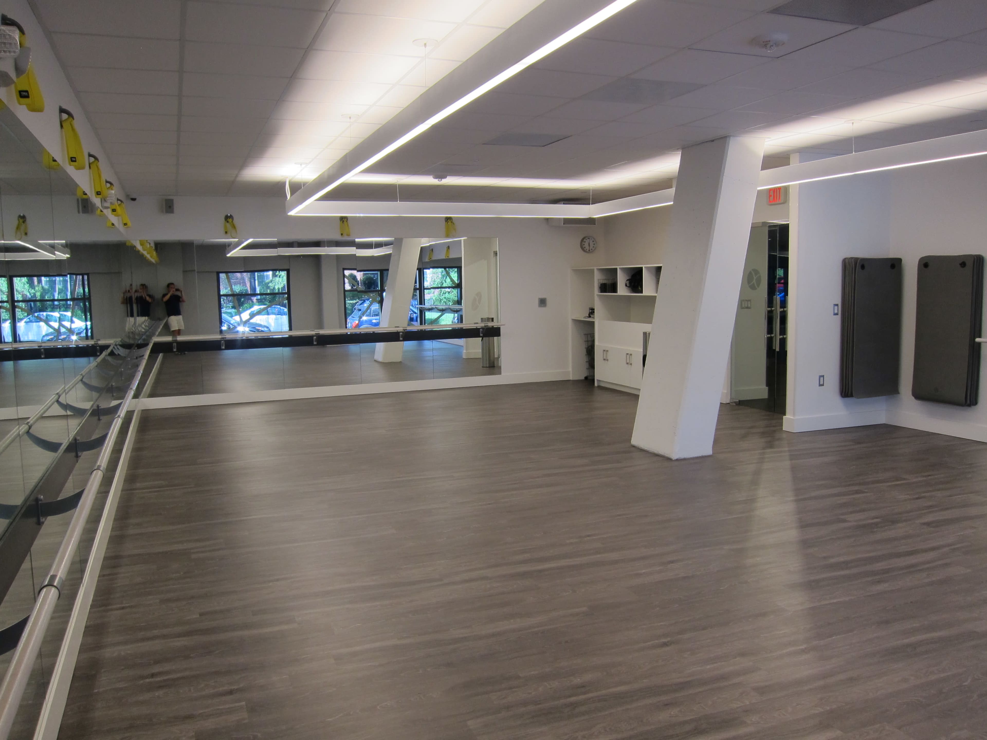 The image shows a spacious, well-lit exercise studio featuring a mirrored wall, wooden flooring, and a minimalist design with fitness equipment stored neatly.