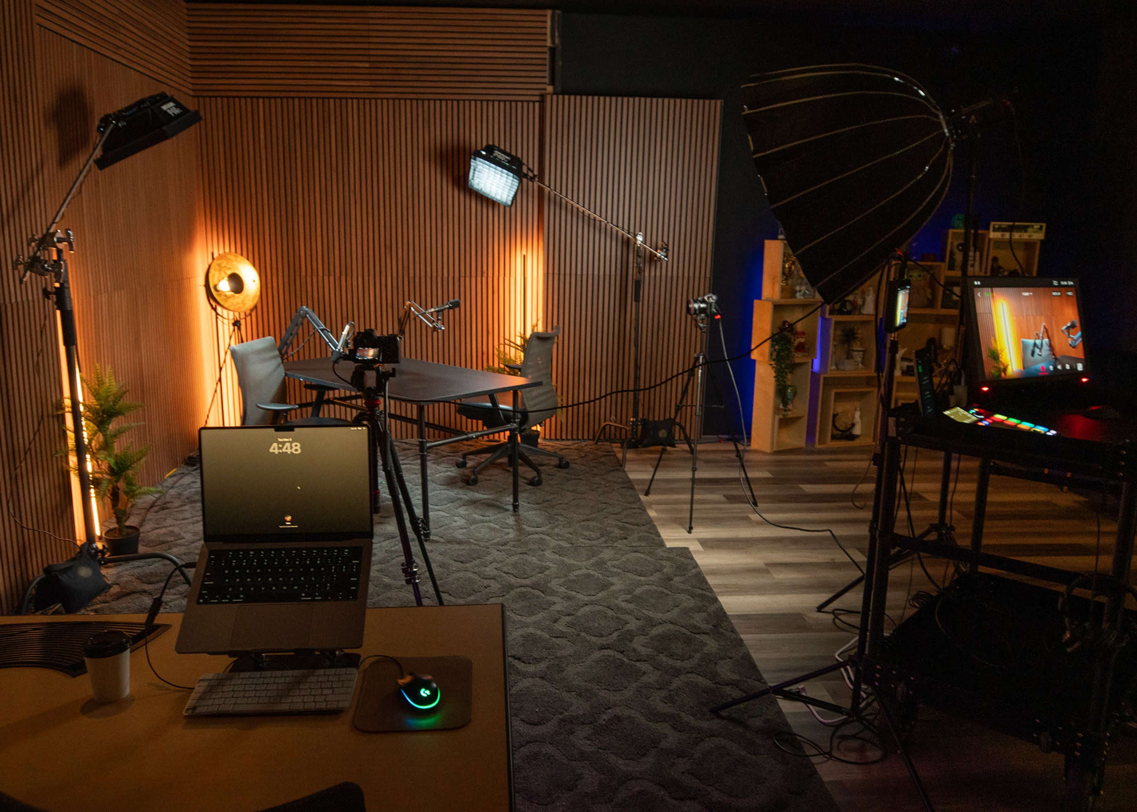 The image shows a modern workspace set up for streaming or recording, featuring a laptop, various lighting equipment, a microphone, and a desk with a chair, all arranged in a warmly lit room with wooden paneling.
