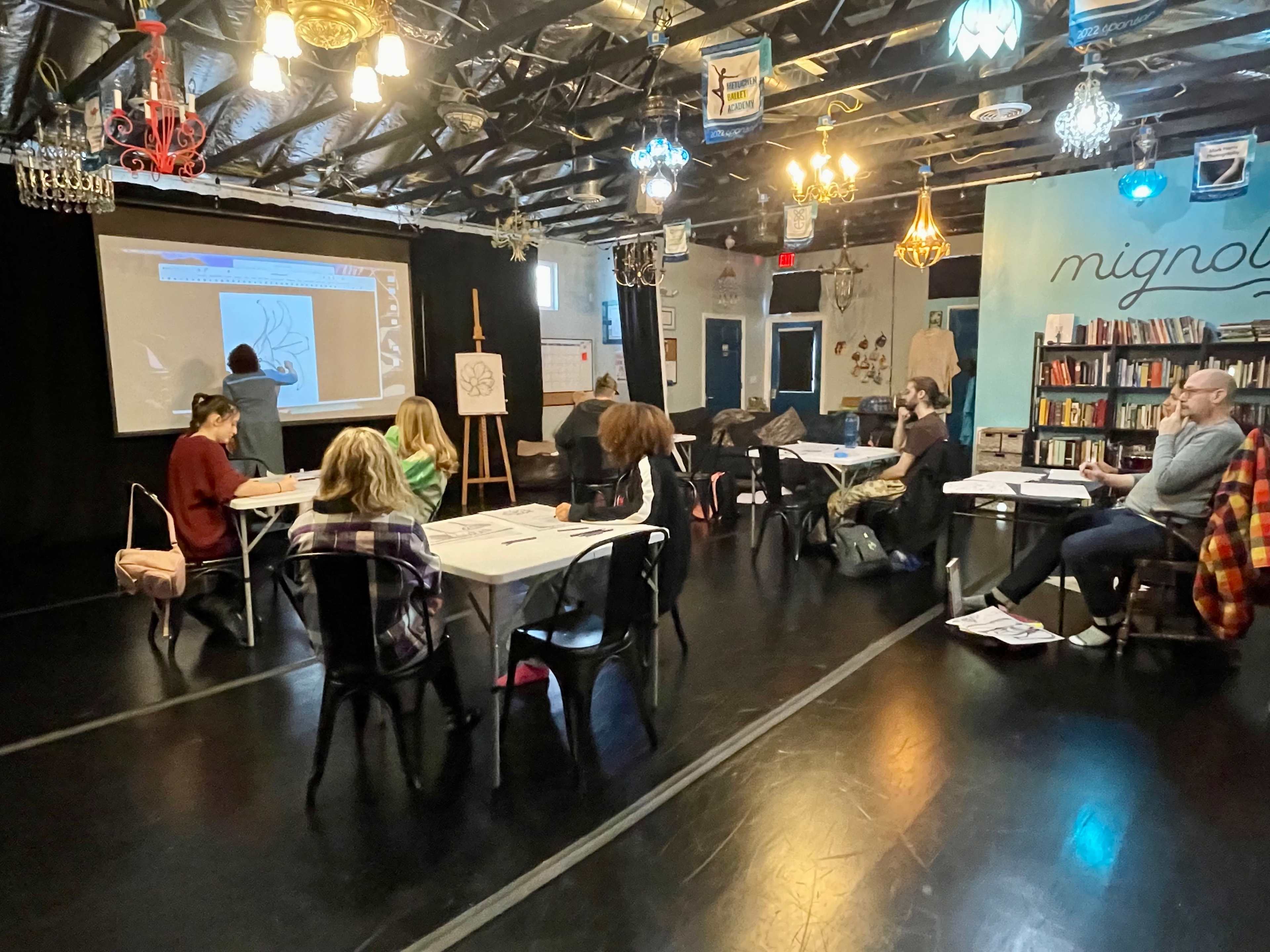 A group of people sits at tables in a studio, watching a presentation projected on a screen while an instructor stands at the front.