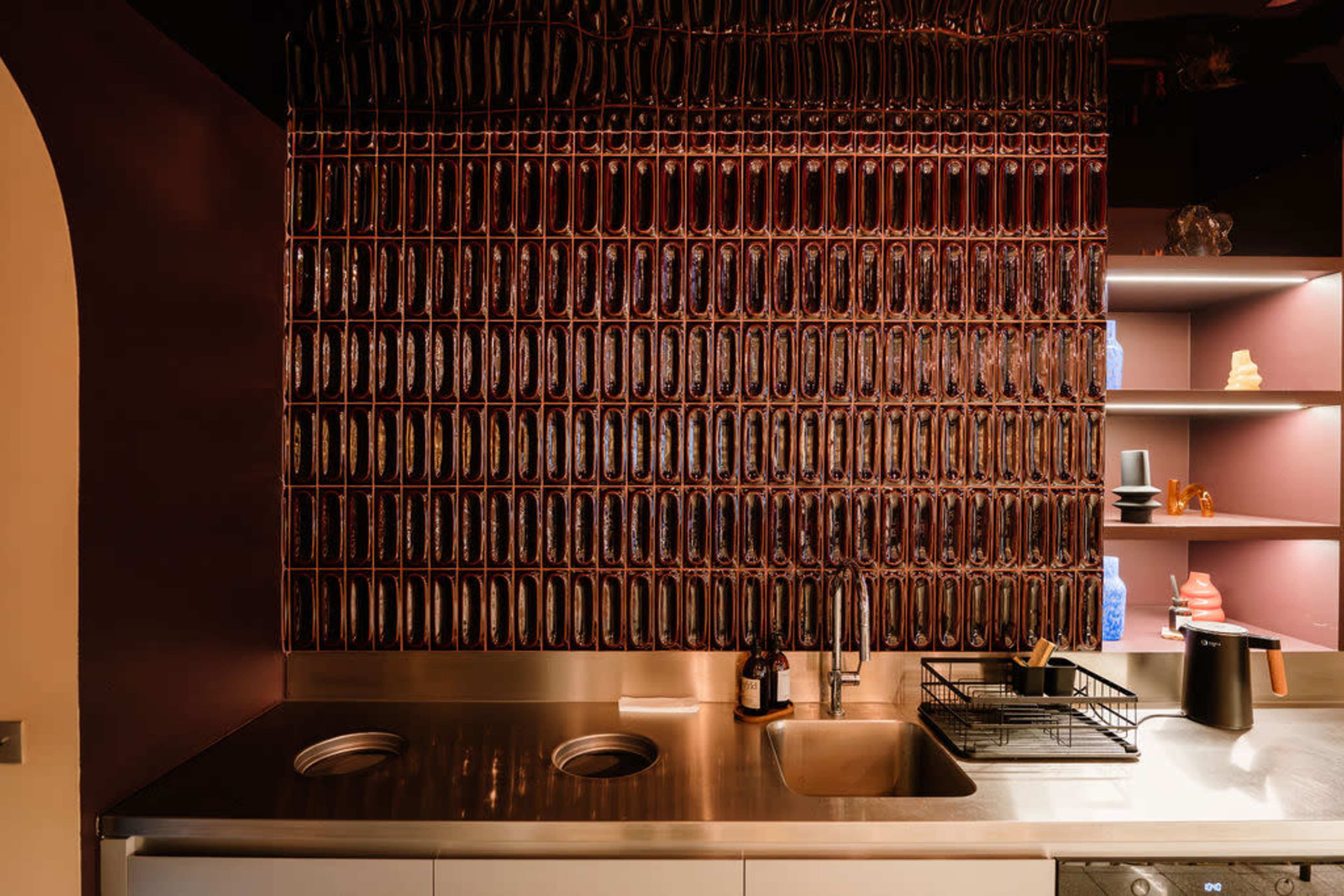 The image features a modern kitchen with a glossy metal countertop, a wall made of patterned glass or plastic tiles, and shelves displaying various kitchen items.