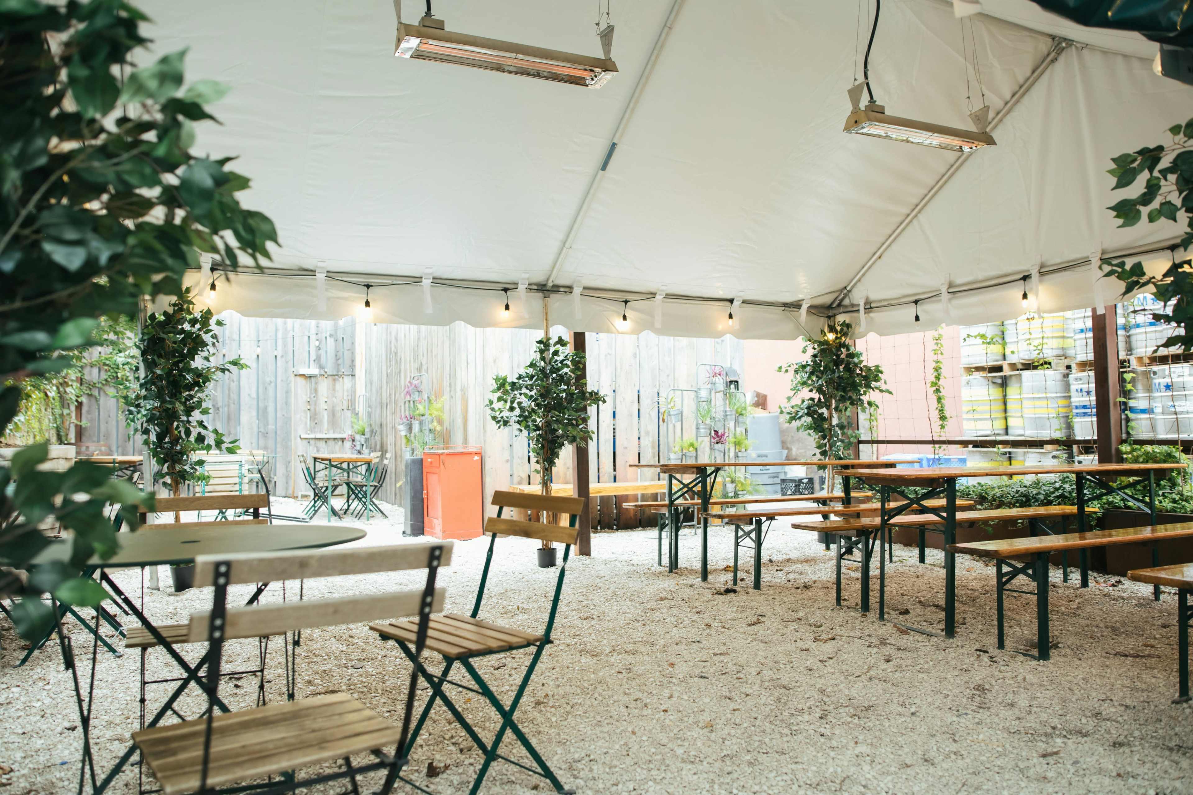 The image shows a spacious outdoor tented area with wooden tables and chairs, surrounded by potted plants and string lights.