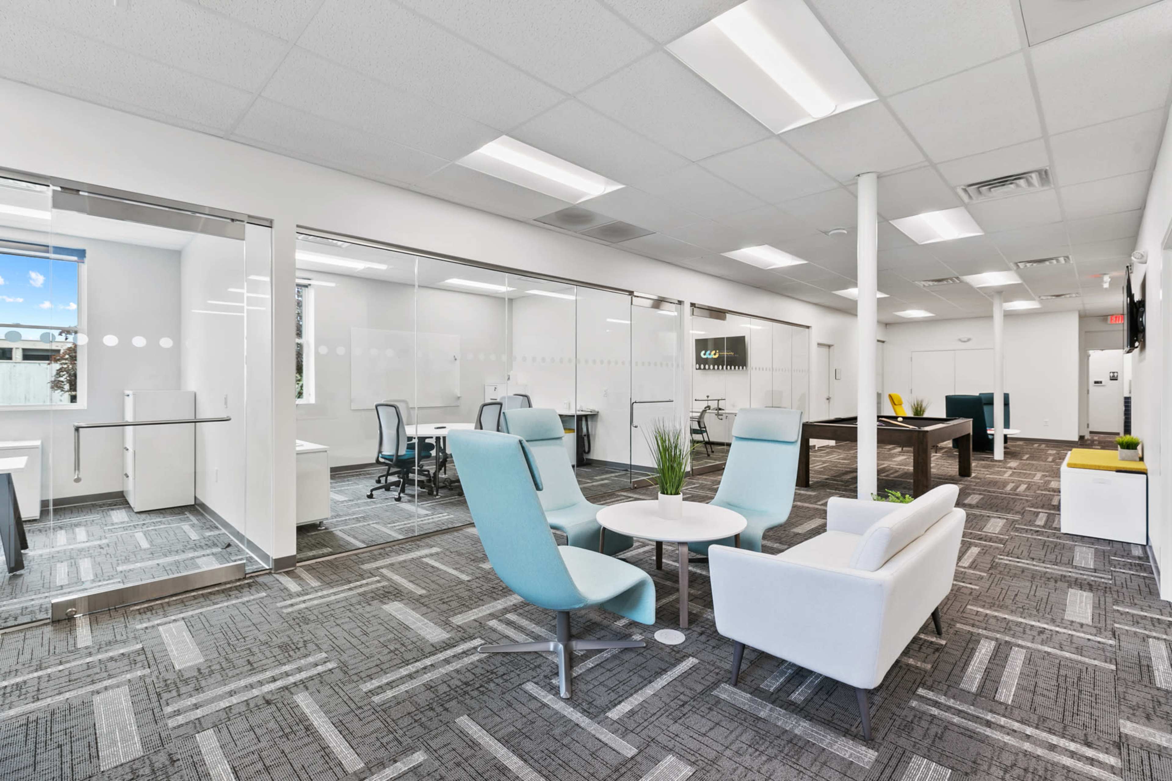 A modern office space features glass-walled meeting rooms, a seating area with blue and white chairs, and a table with chairs.