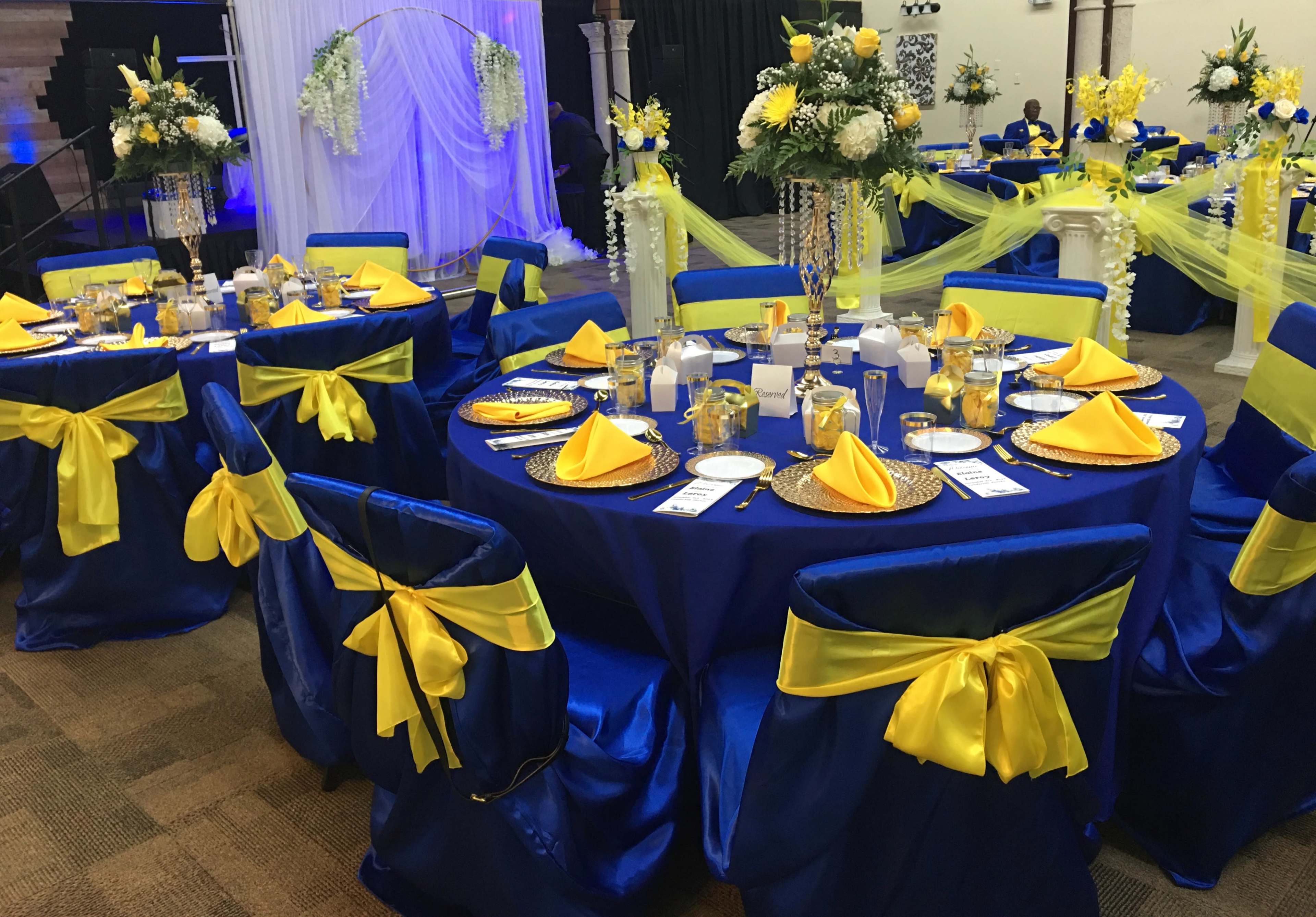 A banquet hall is set with blue and yellow tablecloths, decorated tables with gold chargers, and arrangements of flowers.
