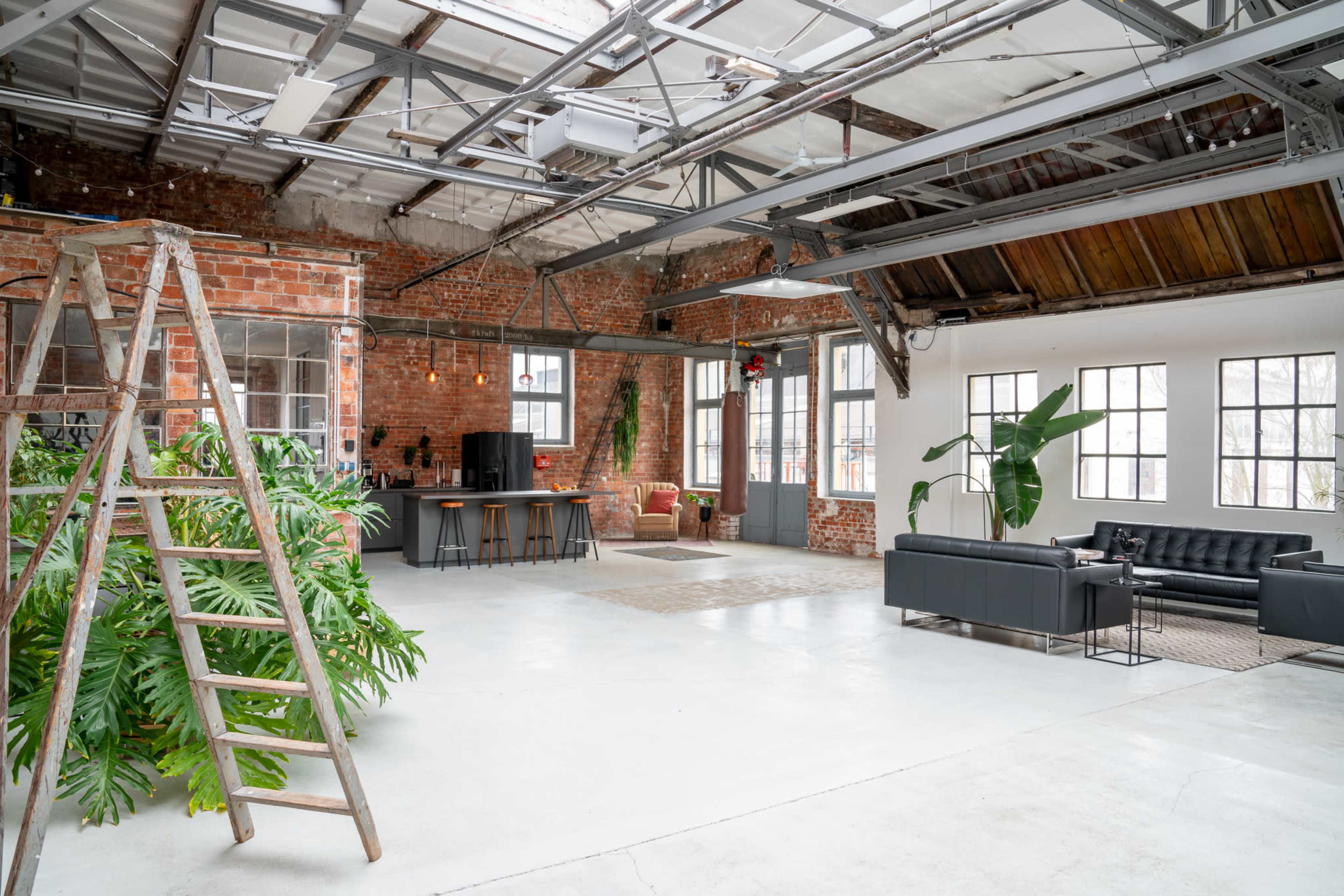 A spacious industrial-style loft with exposed brick walls, large windows, and a mix of green plants, seating areas, and a kitchen.