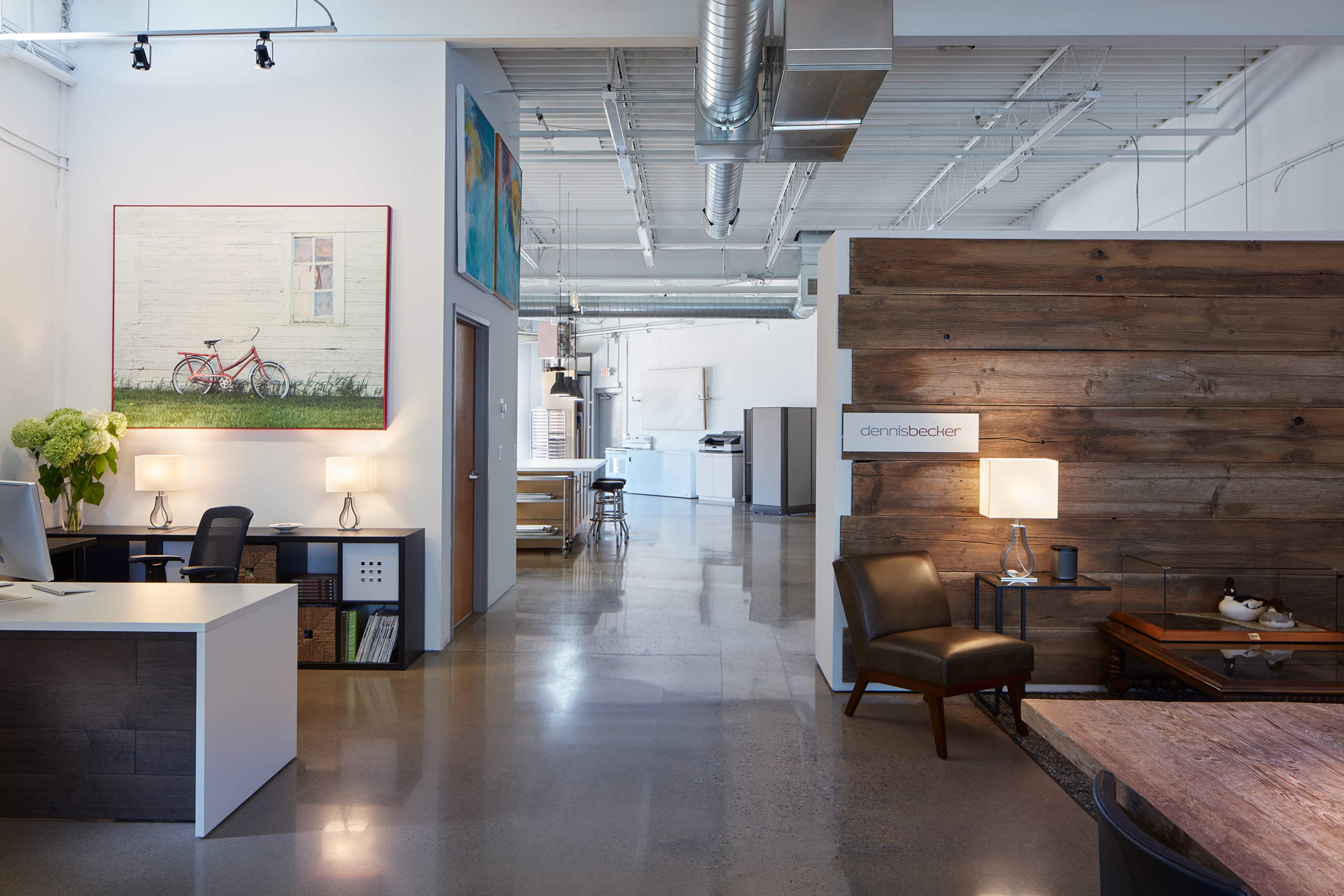 A modern office space with a reception area featuring a desk and artwork, leading into an open layout with wooden accents and a kitchen area in the background.