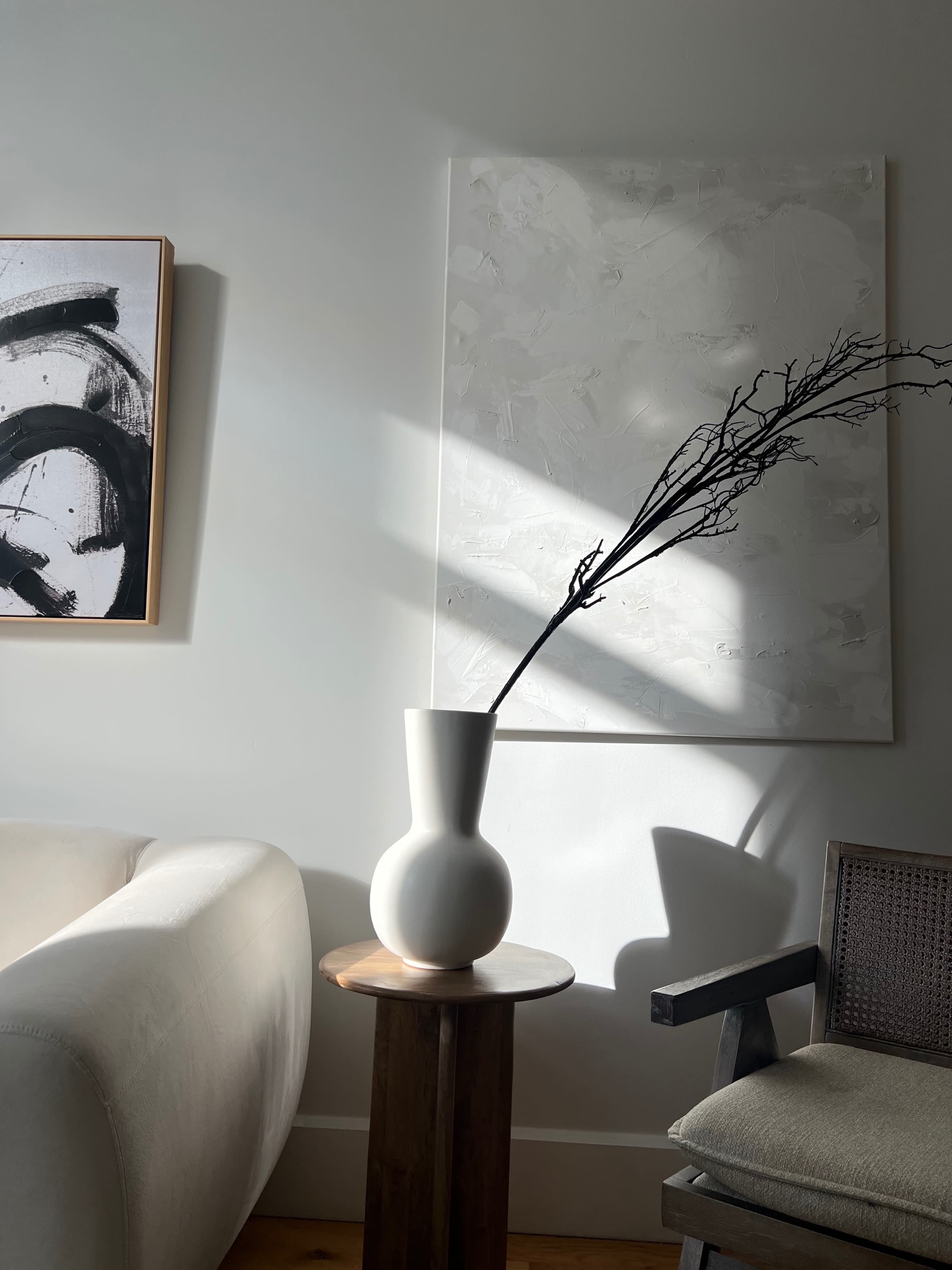 A minimalist interior scene features a white vase with a branch, situated on a wooden side table next to a light-colored chair, against a backdrop of wall art and soft shadows.