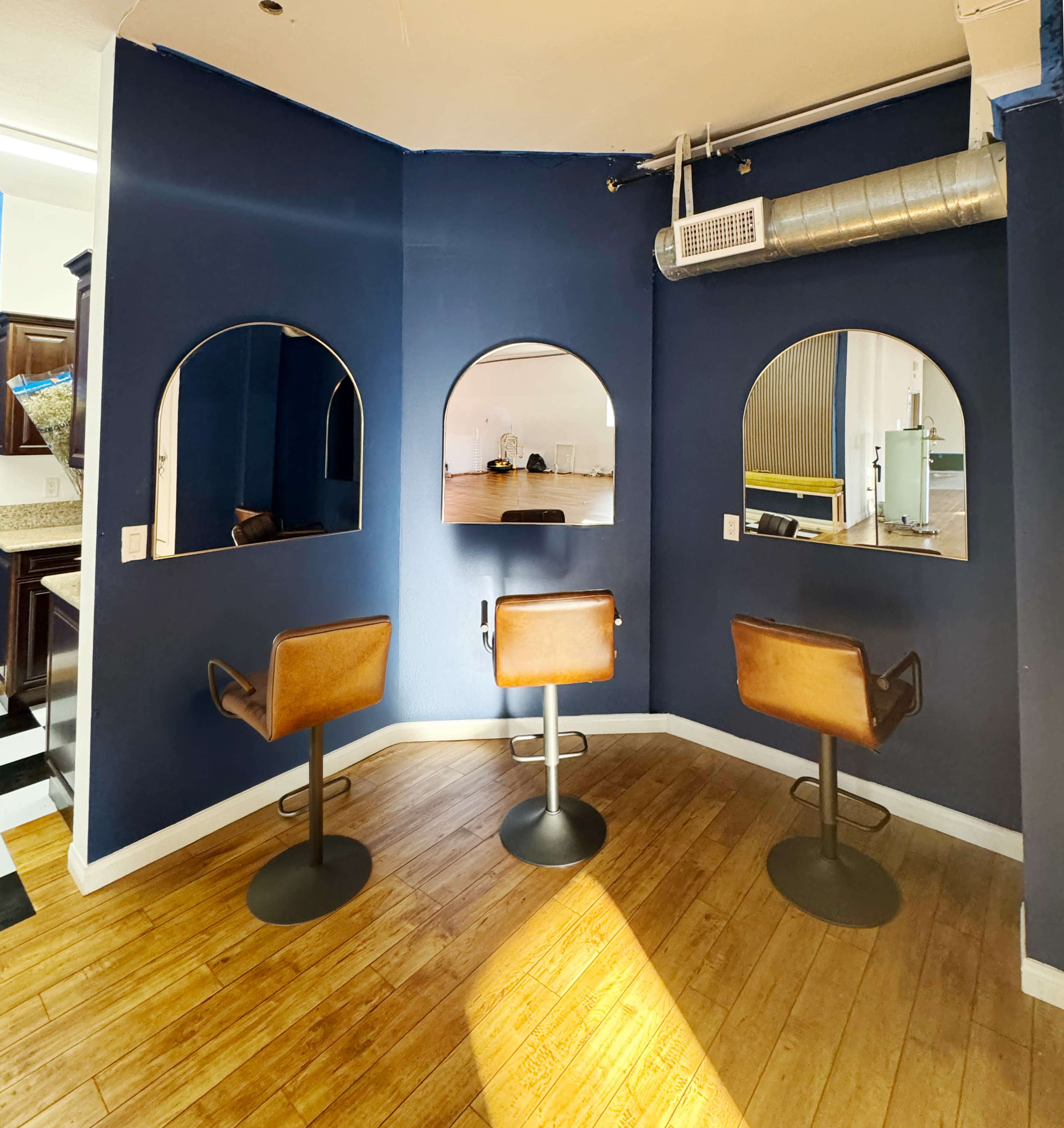 The image shows a corner of a room featuring three brown chairs with metal bases placed in front of arched mirrors against a blue wall.