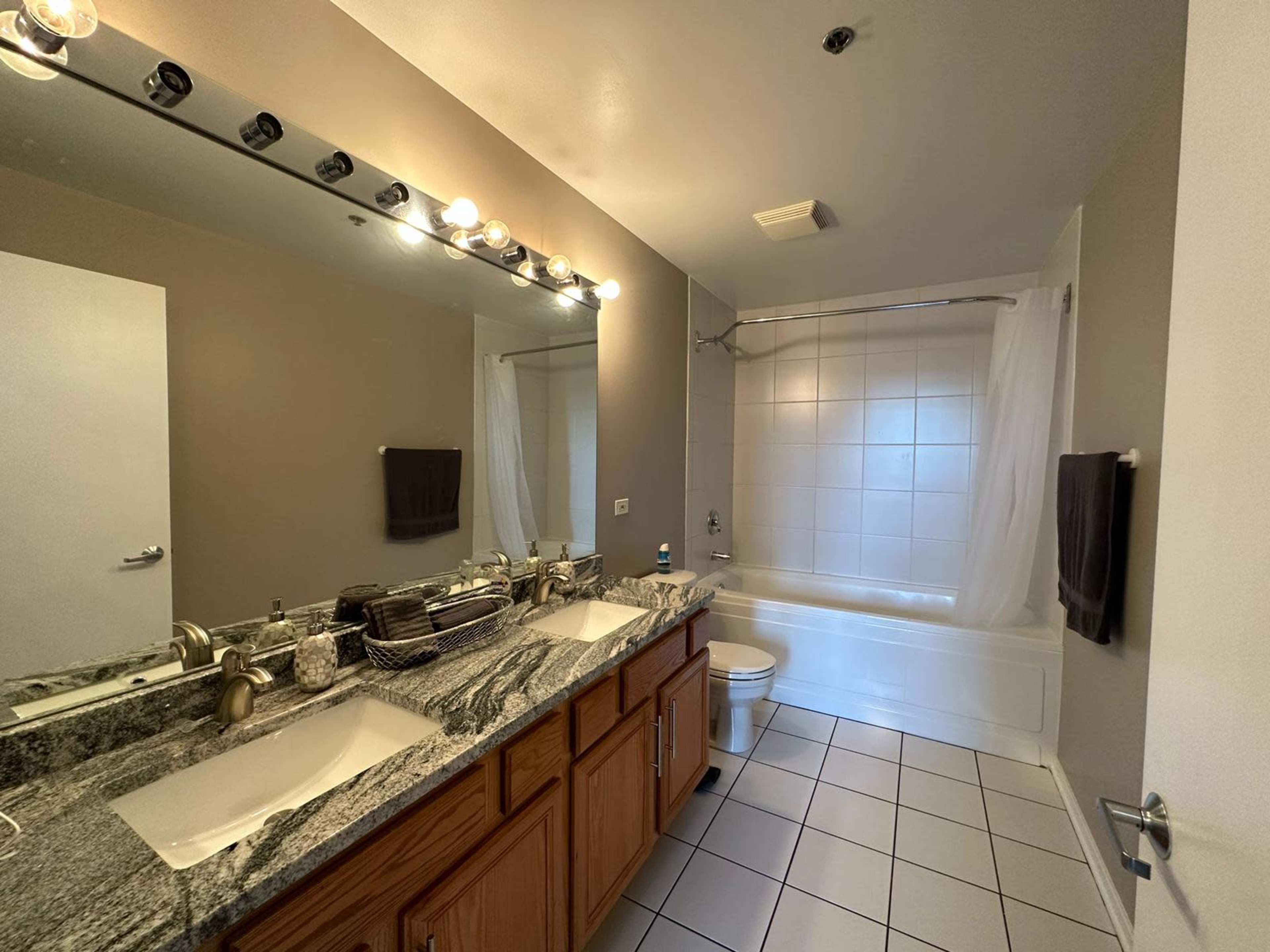 A well-lit bathroom featuring a dual sink vanity with granite countertops, a bathtub with a shower curtain, and light-colored tiled flooring.