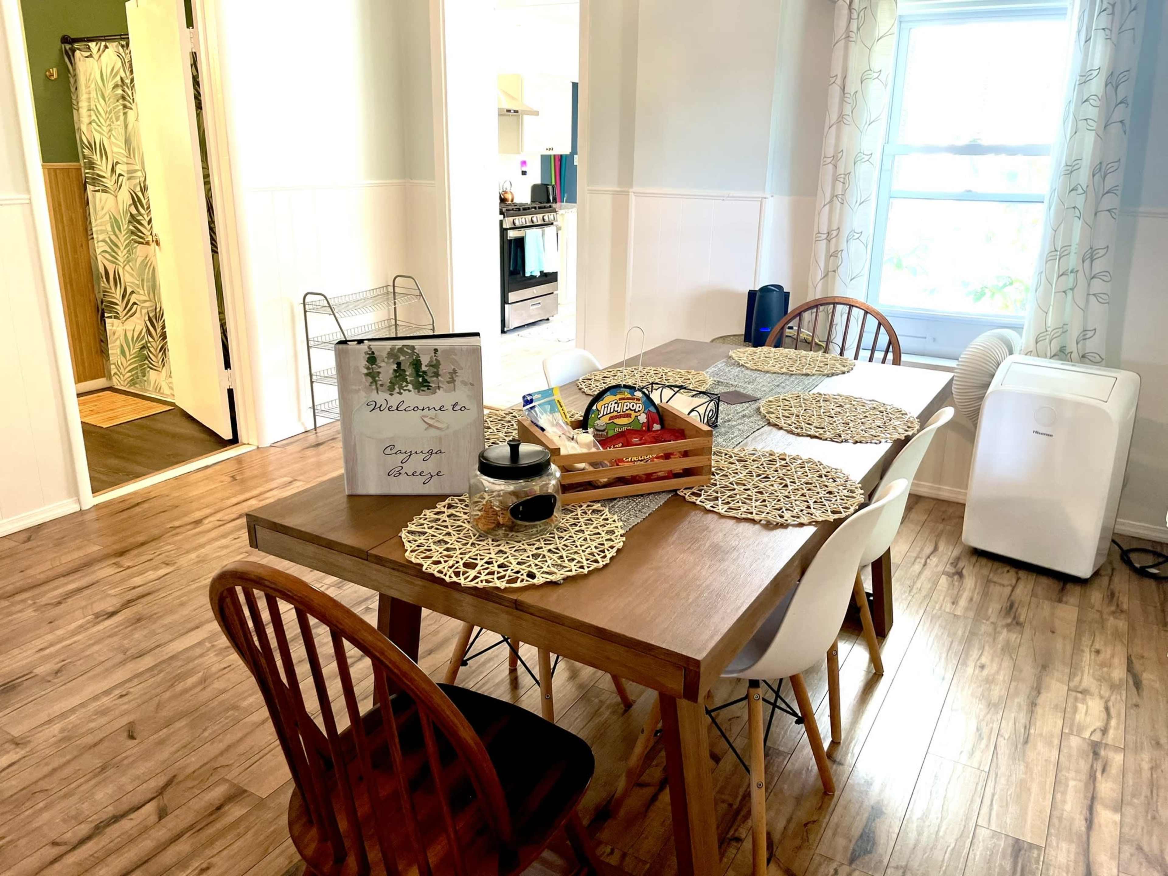 A dining table with placemats and a welcome sign is set in a well-lit room with a kitchen doorway and a fan visible in the background.
