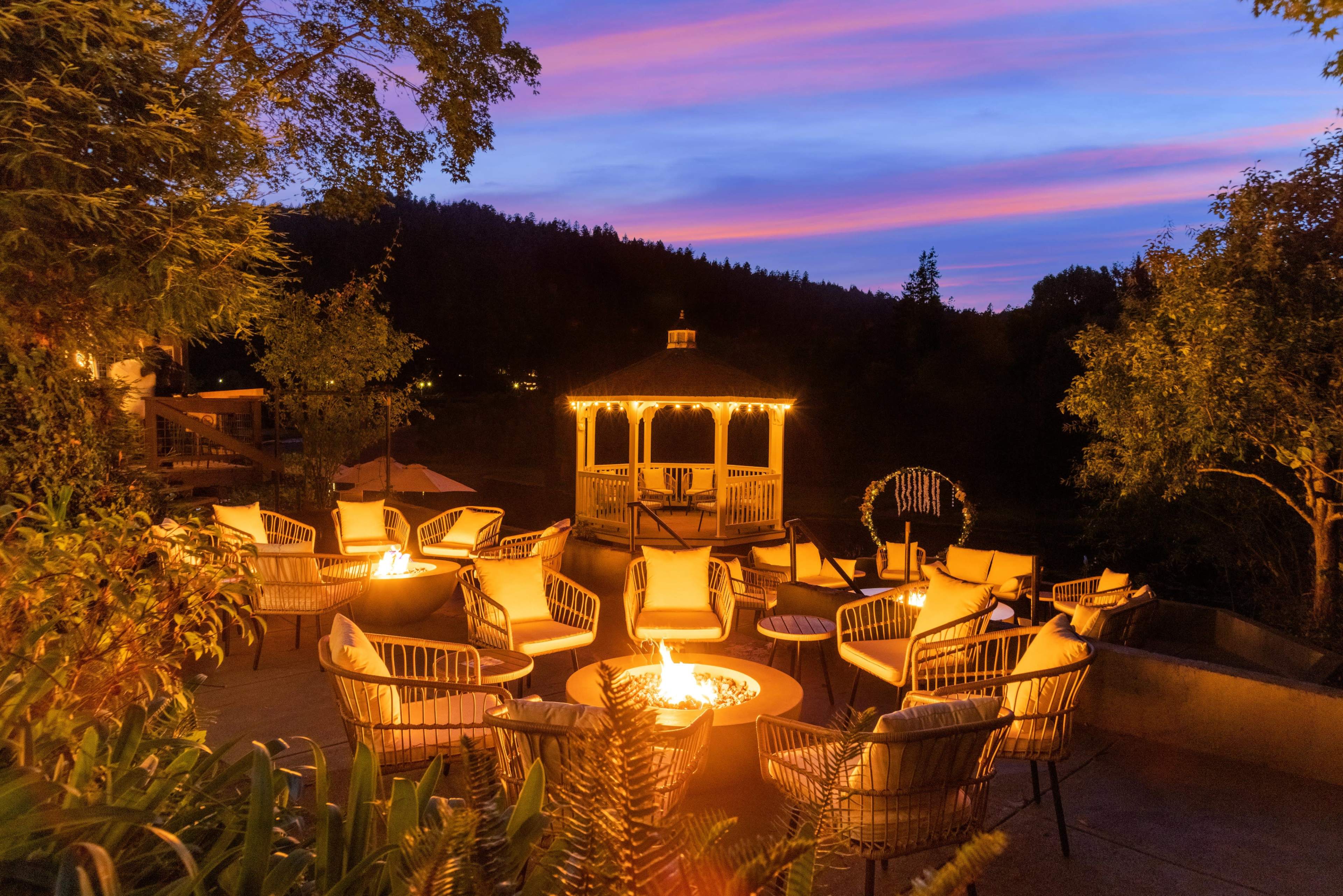 A cozy outdoor seating area with illuminated fire pits surrounded by chairs and a gazebo under a sunset sky.