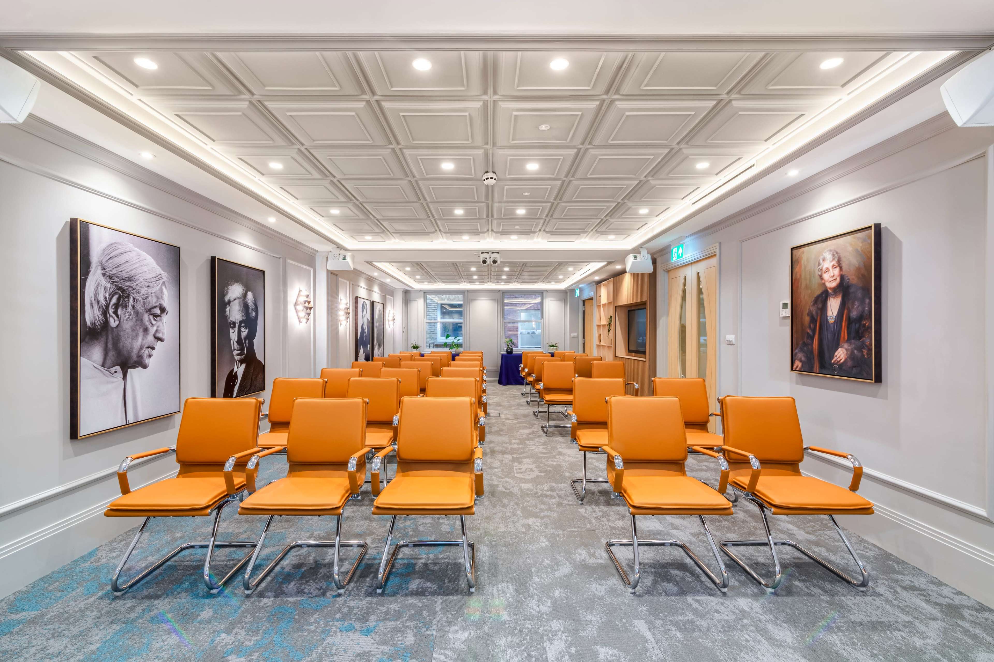 A conference room features rows of orange chairs facing a wall adorned with black-and-white portraits.