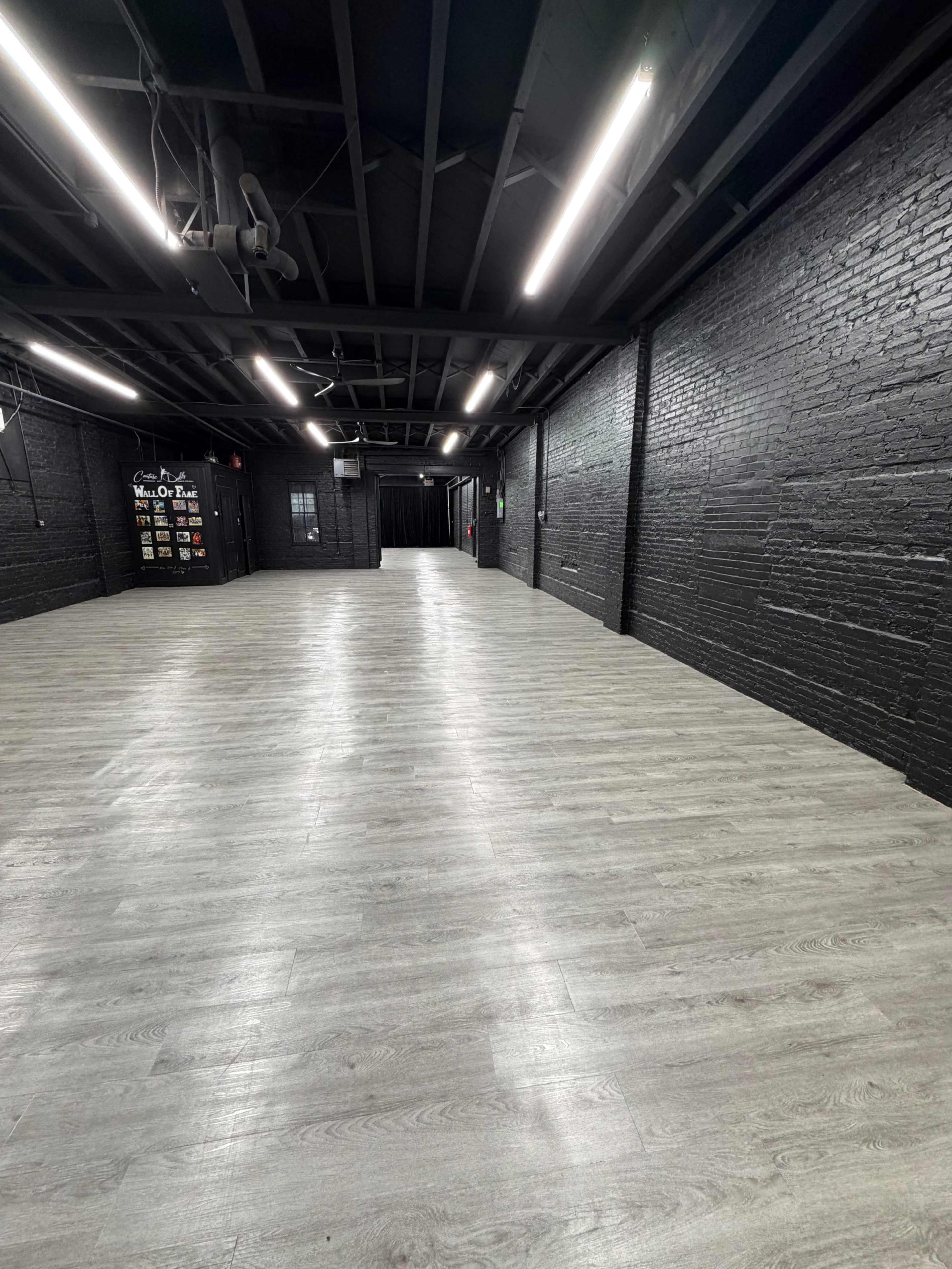 The image shows a spacious, empty industrial-style room with black brick walls and light-colored laminate flooring, illuminated by bright ceiling lights.