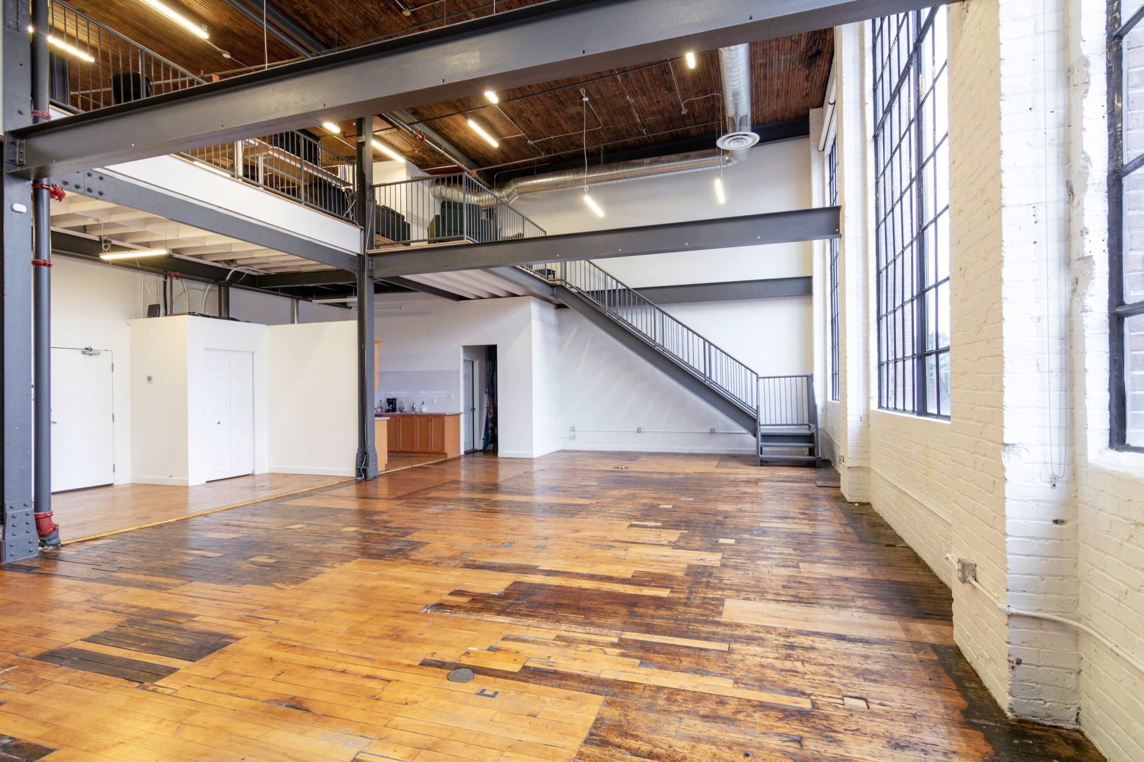 The image shows a spacious, industrial-style loft with wooden floors, large windows, a staircase, and minimalist decor.