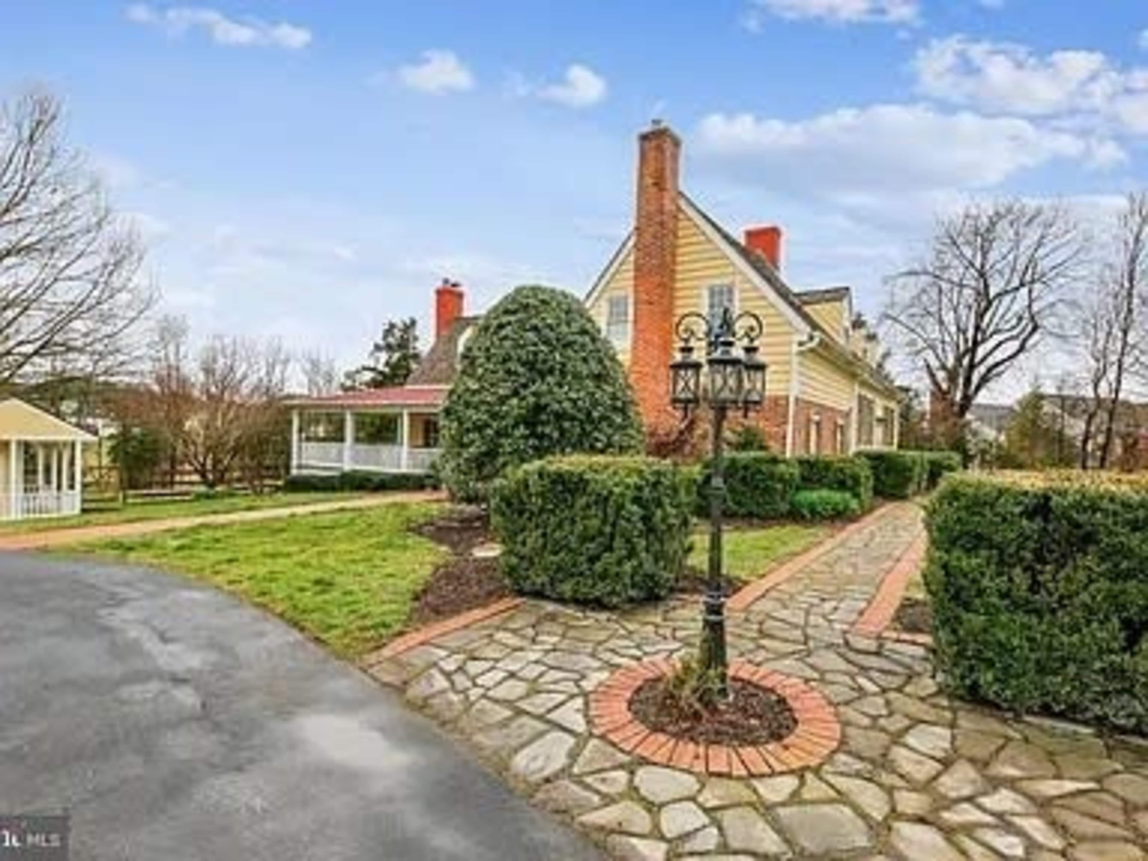 A stone pathway leads to a large house with a sloped roof, surrounded by manicured shrubs and trees.