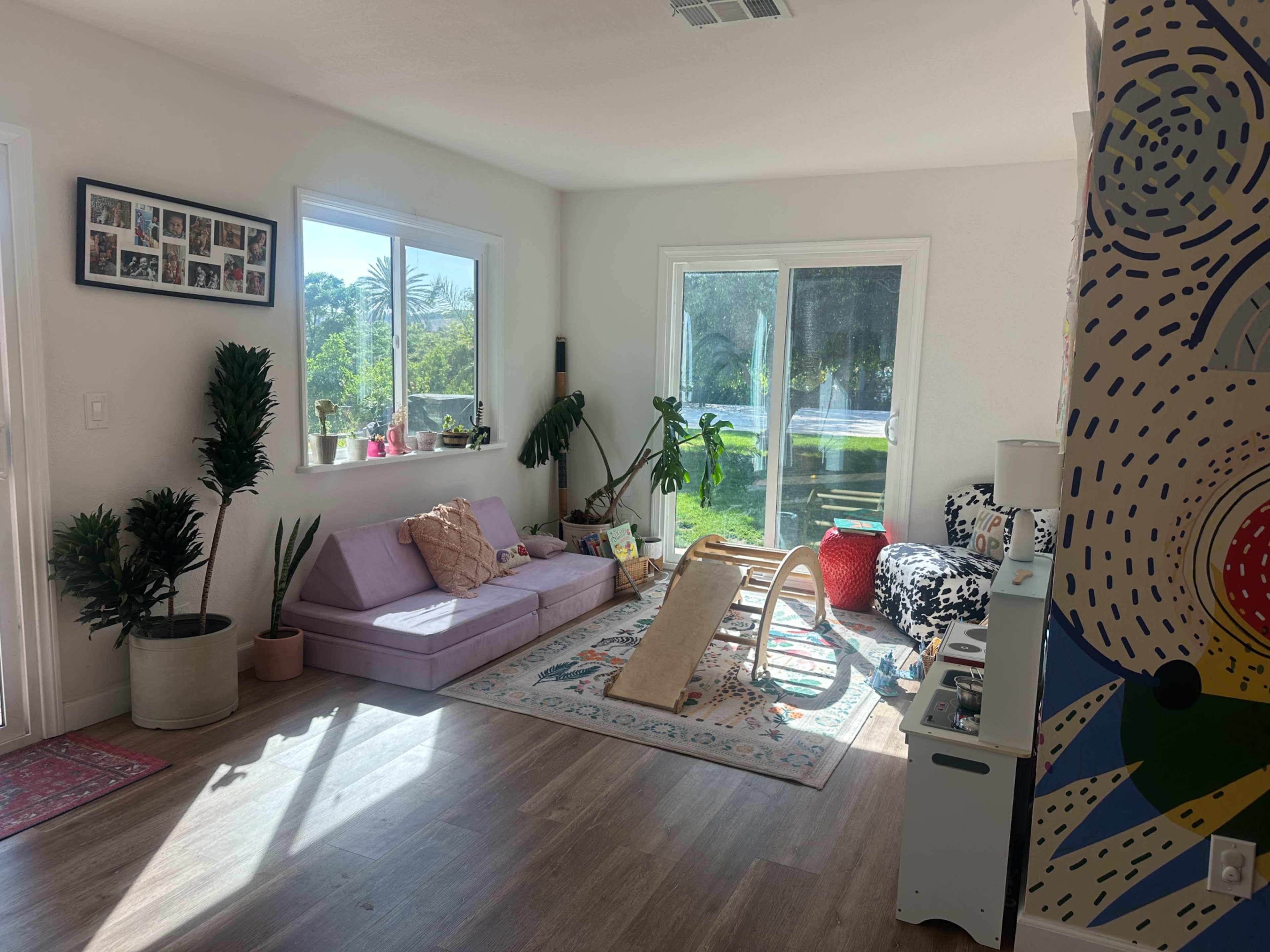 The image shows a bright living room featuring a purple sofa, a rug, various green plants, and colorful decor elements near large windows.