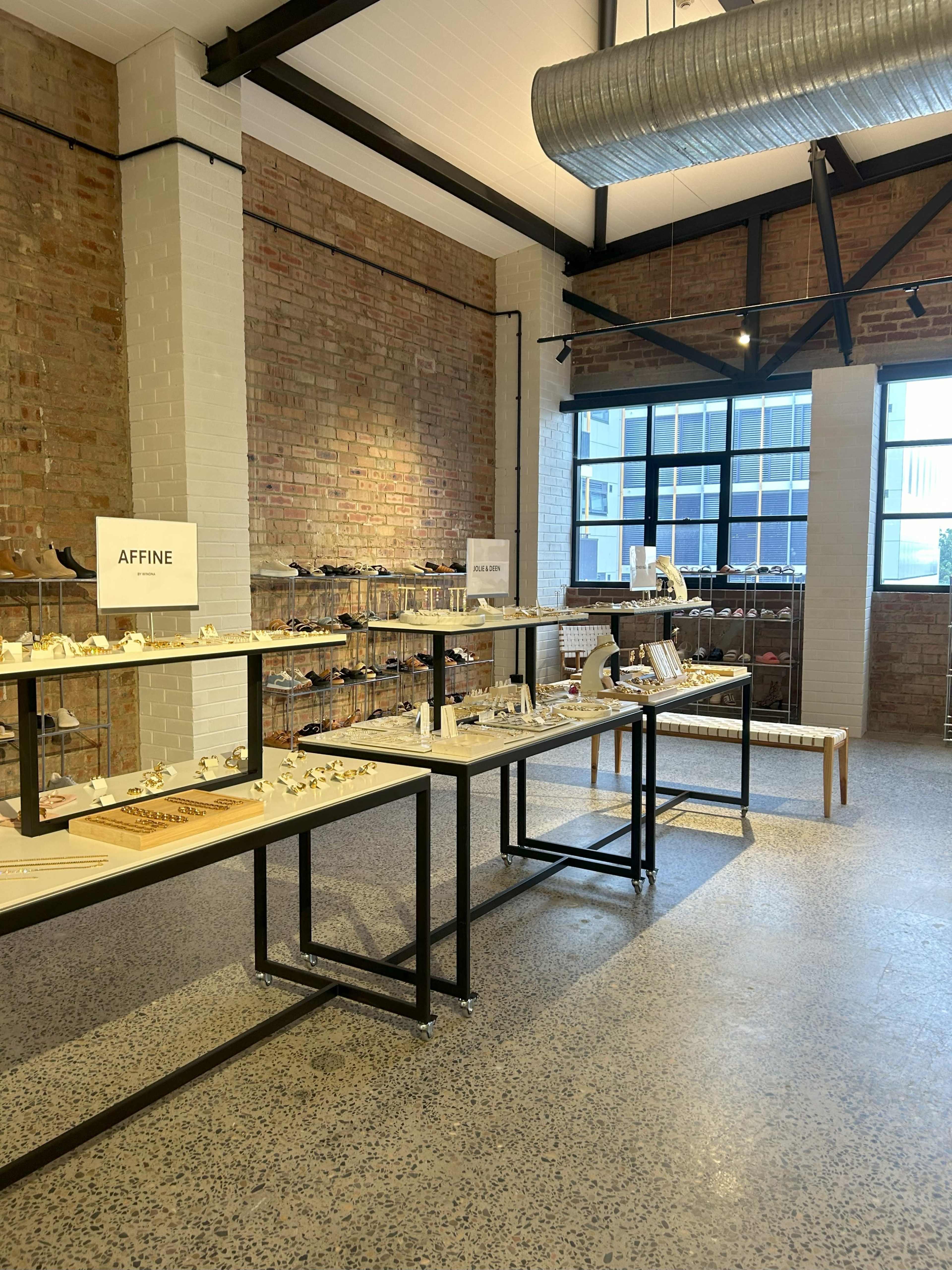 A bright, modern retail space featuring tables displaying various jewelry items against a backdrop of exposed brick walls and large windows.