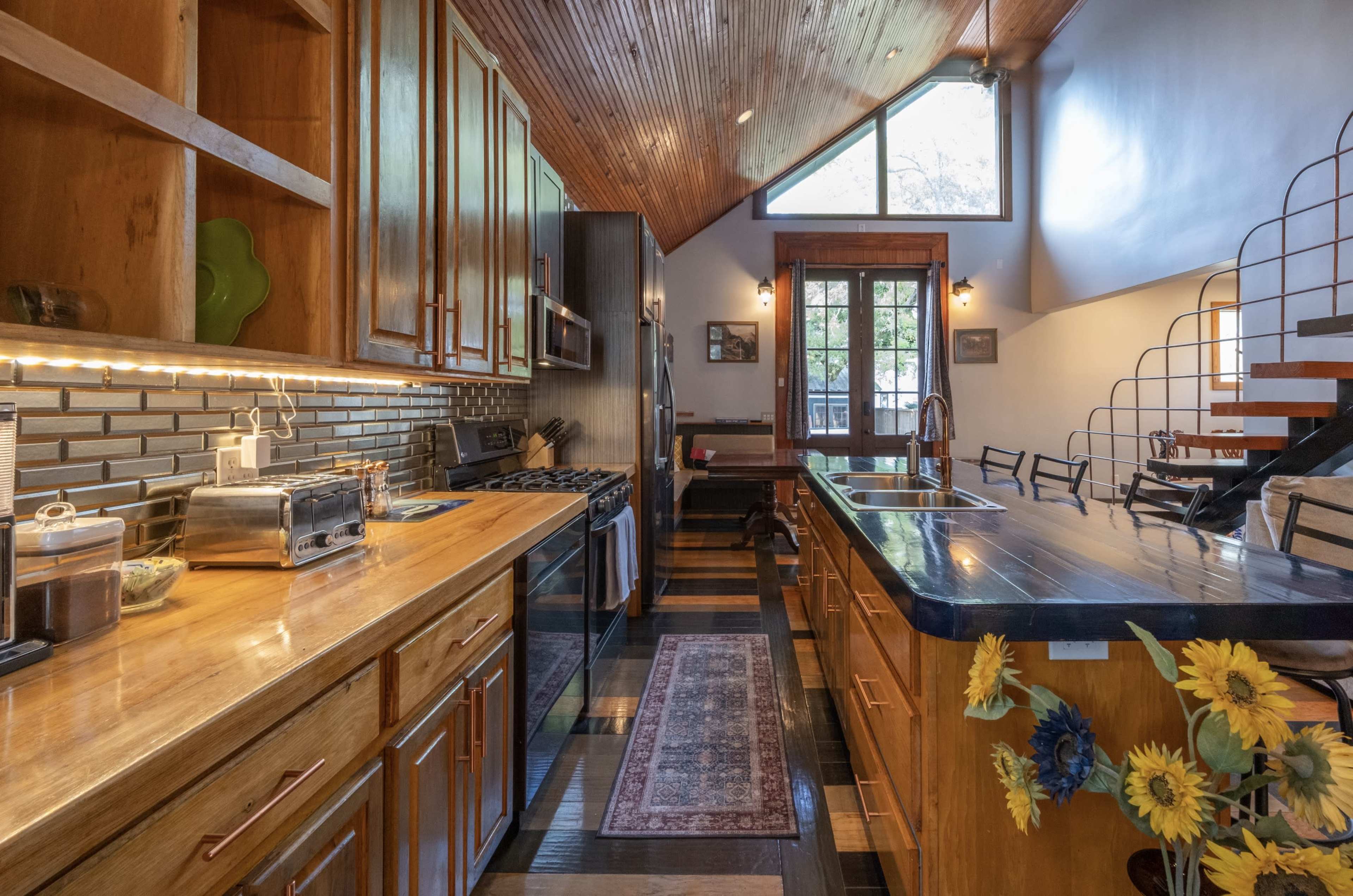 The image shows a modern kitchen with wooden cabinetry, a central island with a dark countertop, and large windows allowing natural light to enter the space.