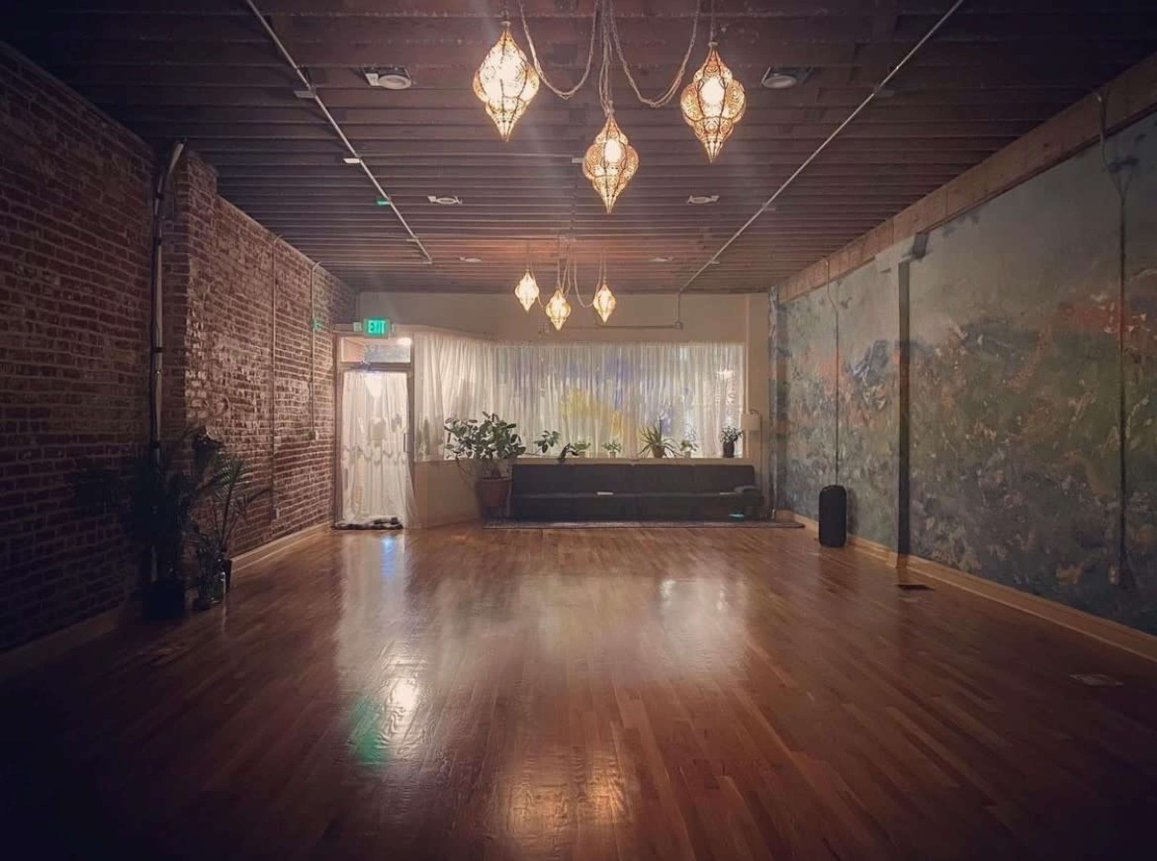 A spacious, well-lit room features hardwood floors, exposed brick walls, and decorative hanging lights, with a mural and plants along one side.