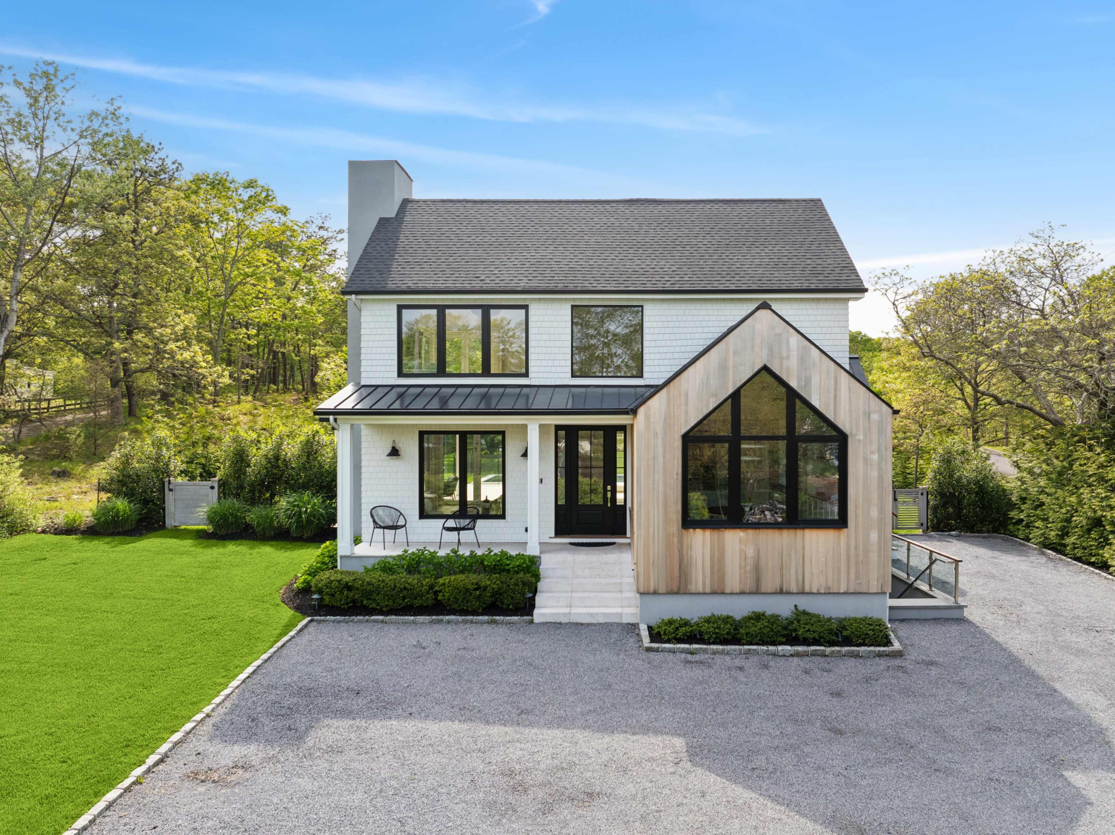The image shows a modern two-story house featuring a mix of white siding and wood accents, set on a landscaped lot with a green lawn and gravel driveway.