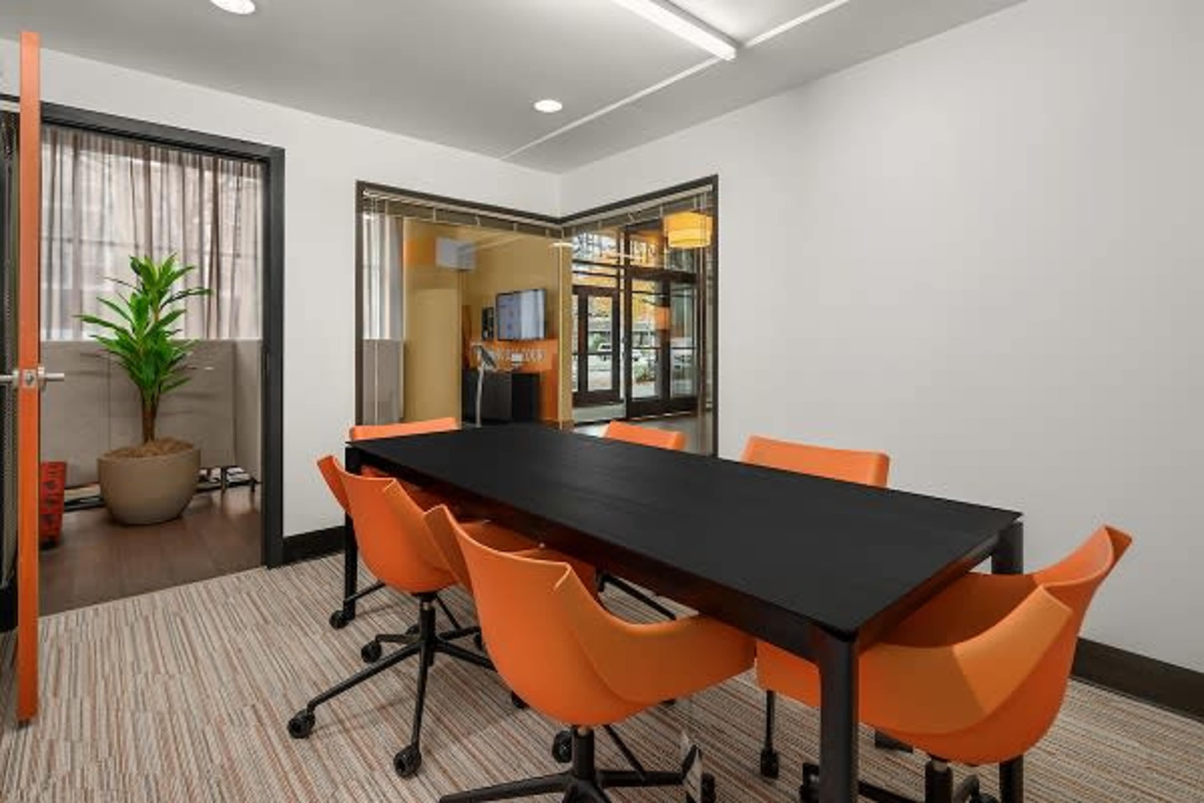 A conference room features a black table surrounded by six orange chairs, with a plant in a pot visible in the background.