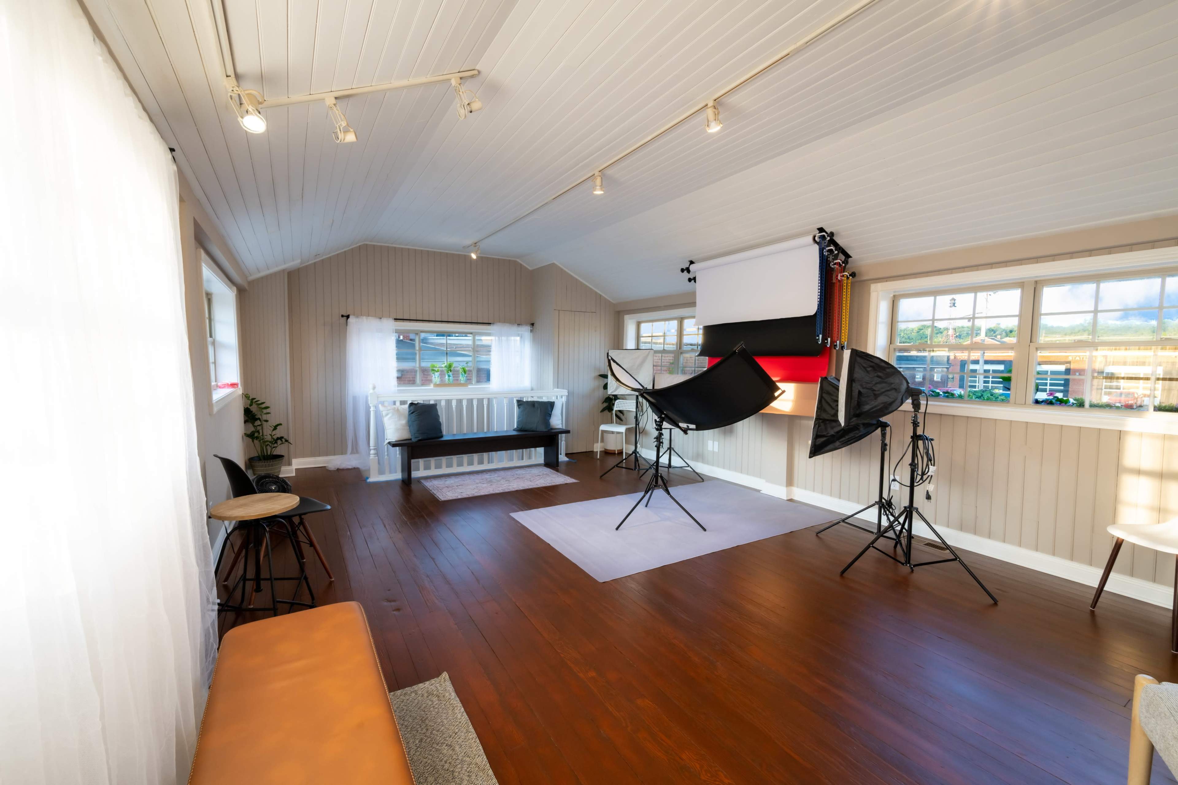 The image shows a well-lit interior photography studio with a wooden floor, white walls, and lighting equipment set up alongside a seating area.