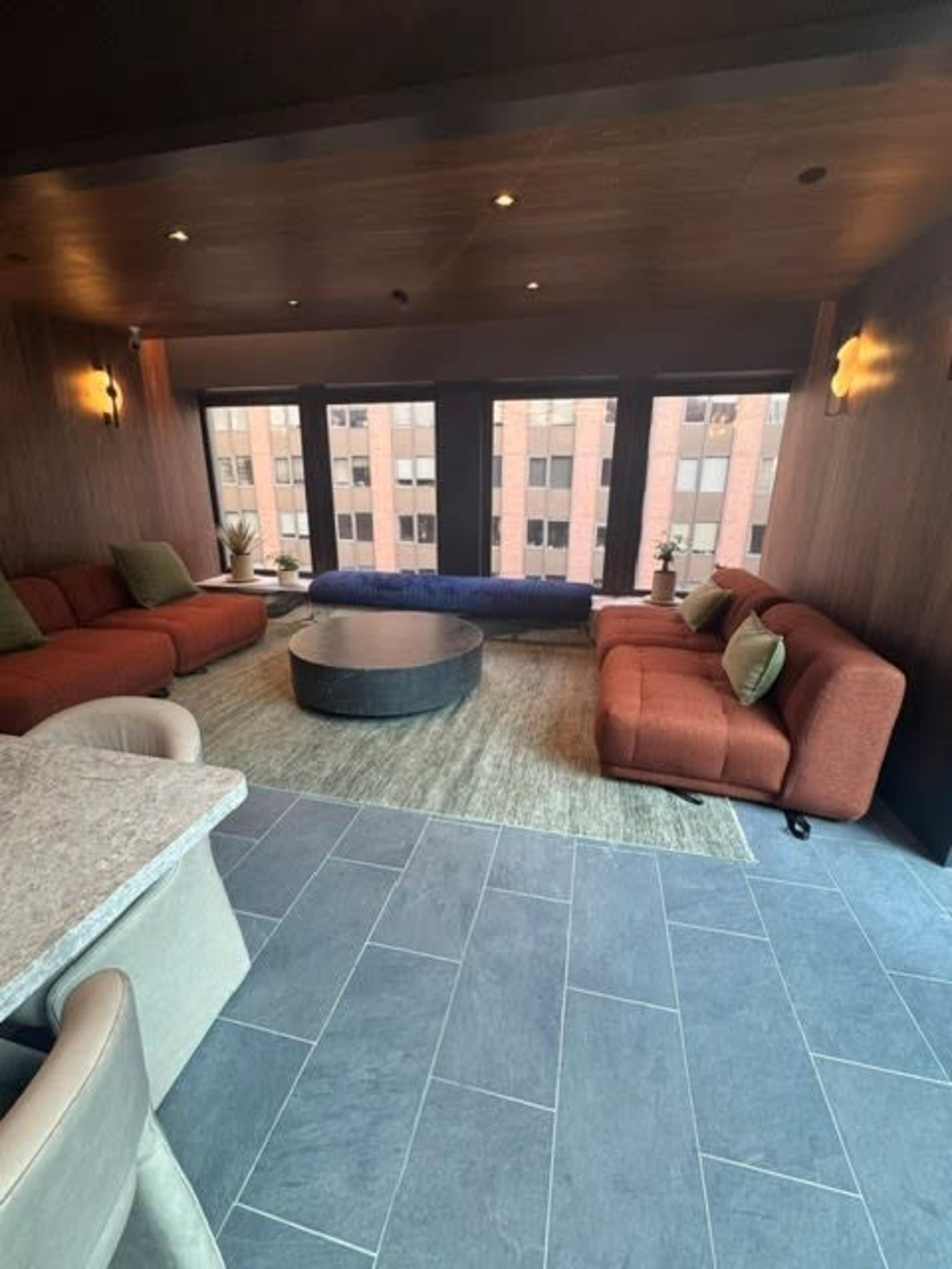 A modern lounge features curved orange sofas, a round coffee table, and large windows offering a view of a cityscape.