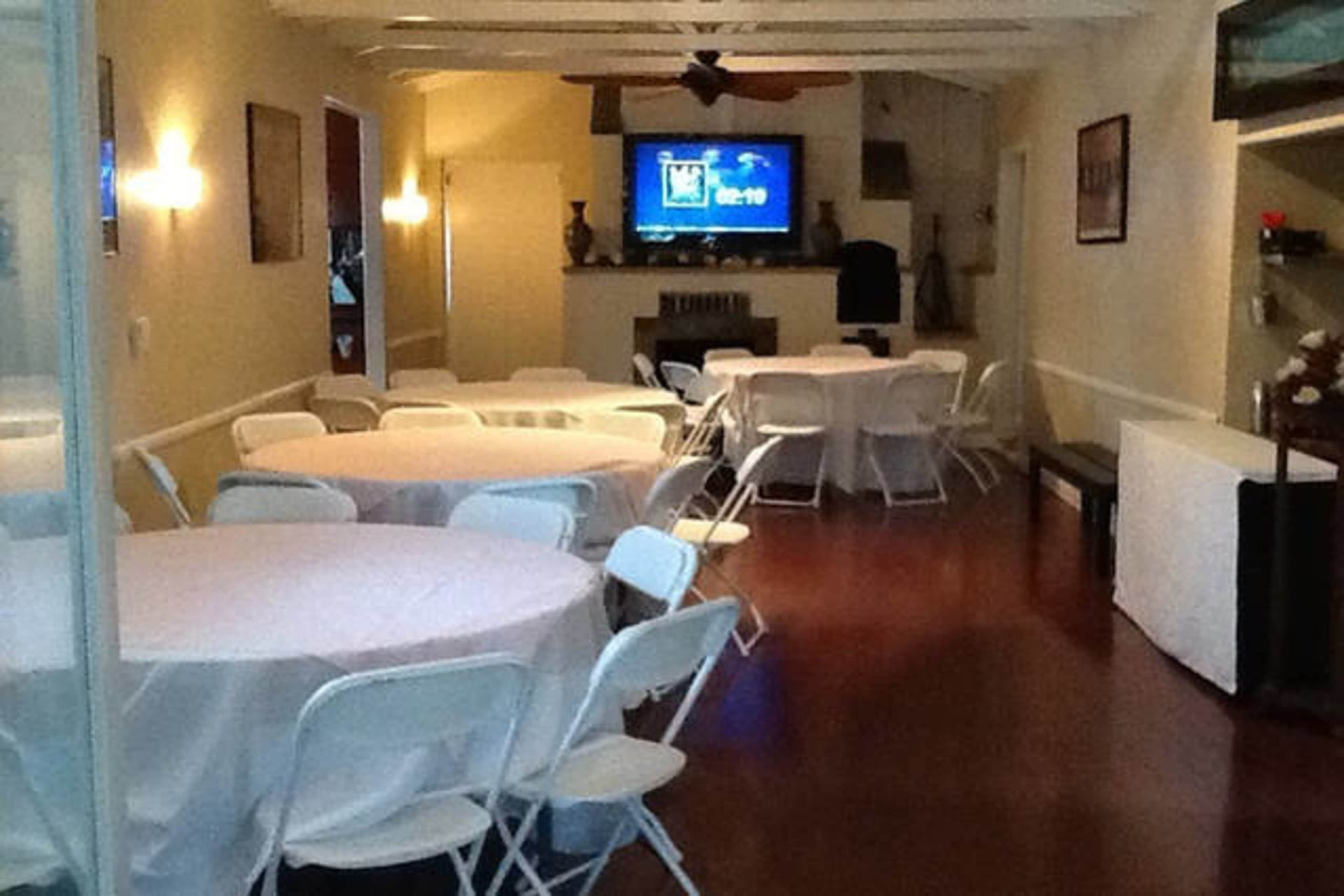 A banquet hall with several round tables set with white tablecloths and a large television screen at the end.