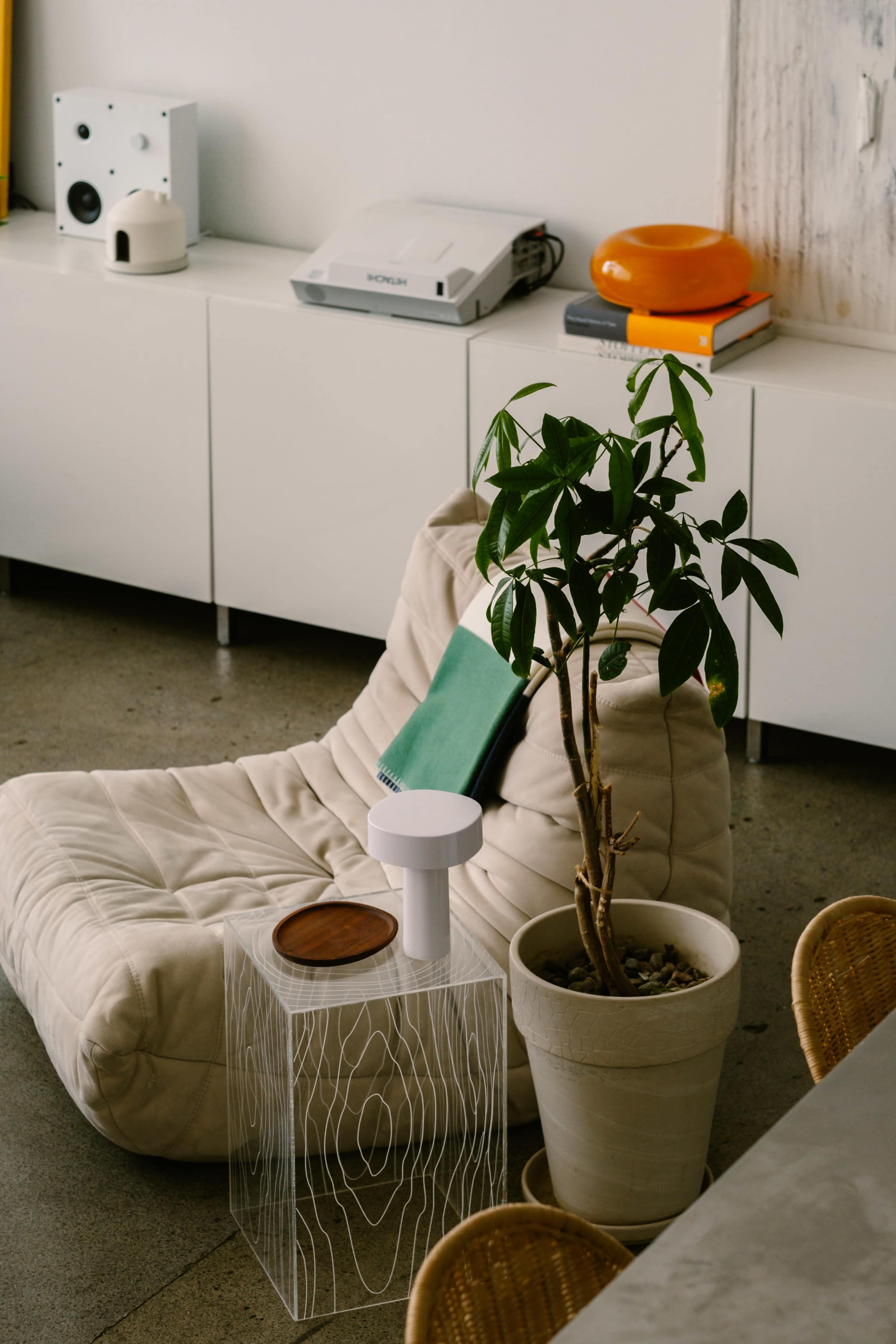 A white, cushioned lounge chair sits next to a small, clear table with a potted plant and a wooden coaster on it, in a modern, well-lit room.