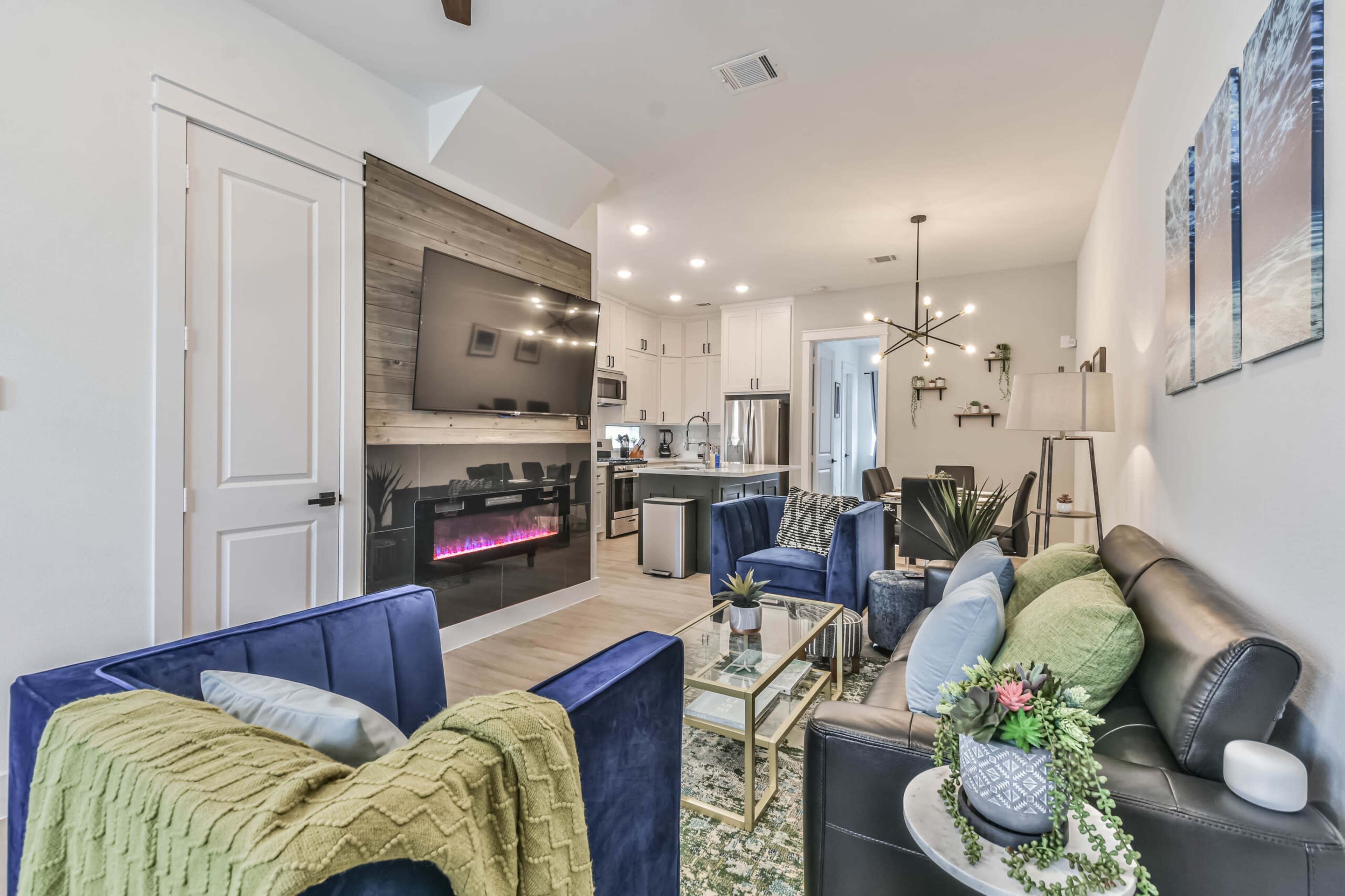 The living room features two sofas, a glass coffee table, and a sleek electric fireplace against a modern backdrop of open kitchen space.