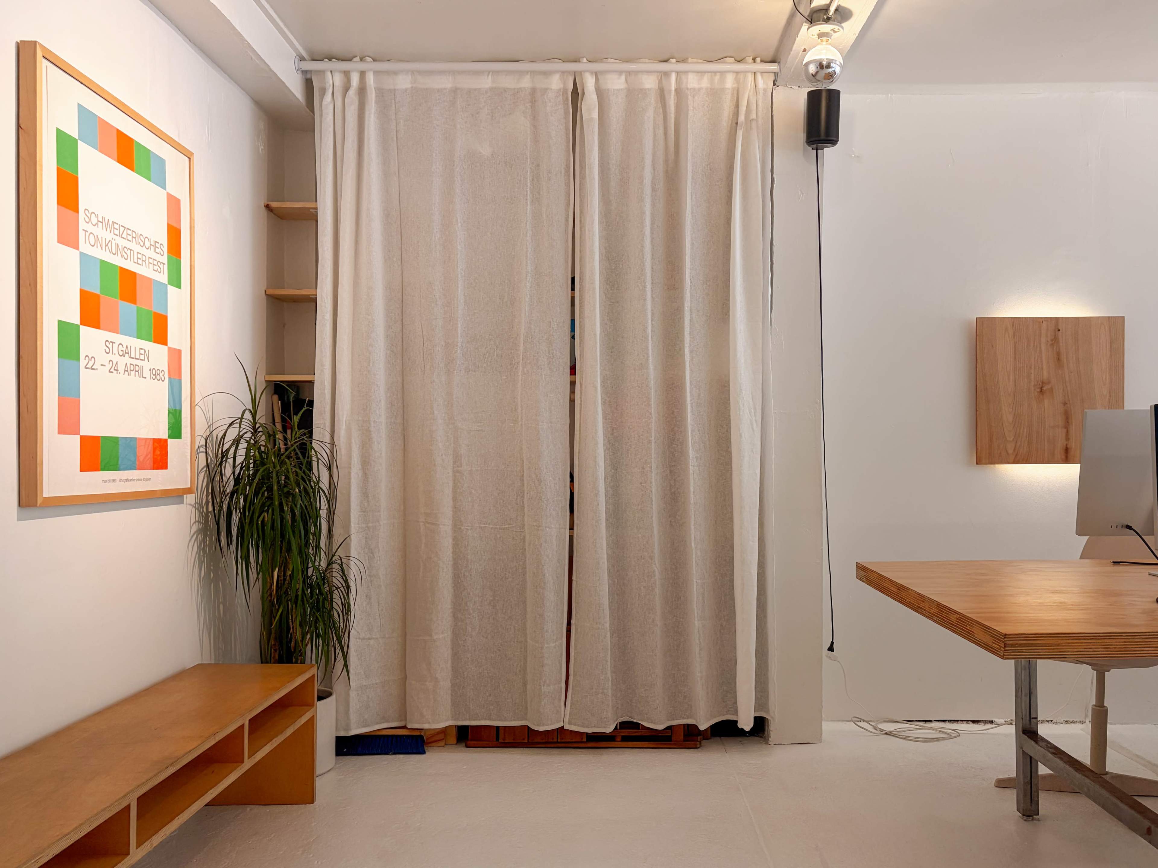 The image shows a minimalist room featuring a wooden table, a plant, and curtains that partially hide a space behind them, with artwork and a wall-mounted panel visible.