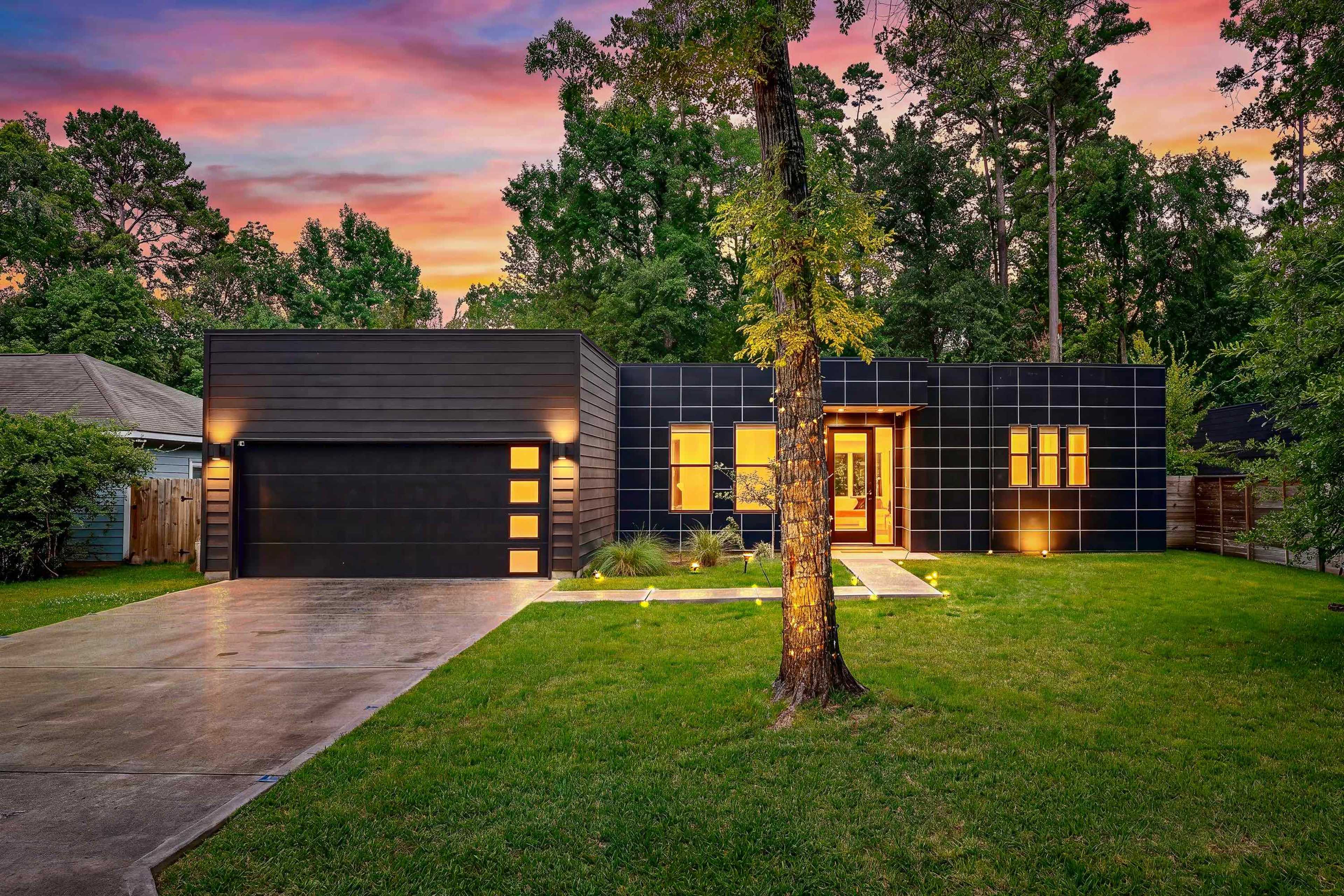A modern house with a black exterior and large windows is set against a colorful sunset, surrounded by trees and a green lawn.