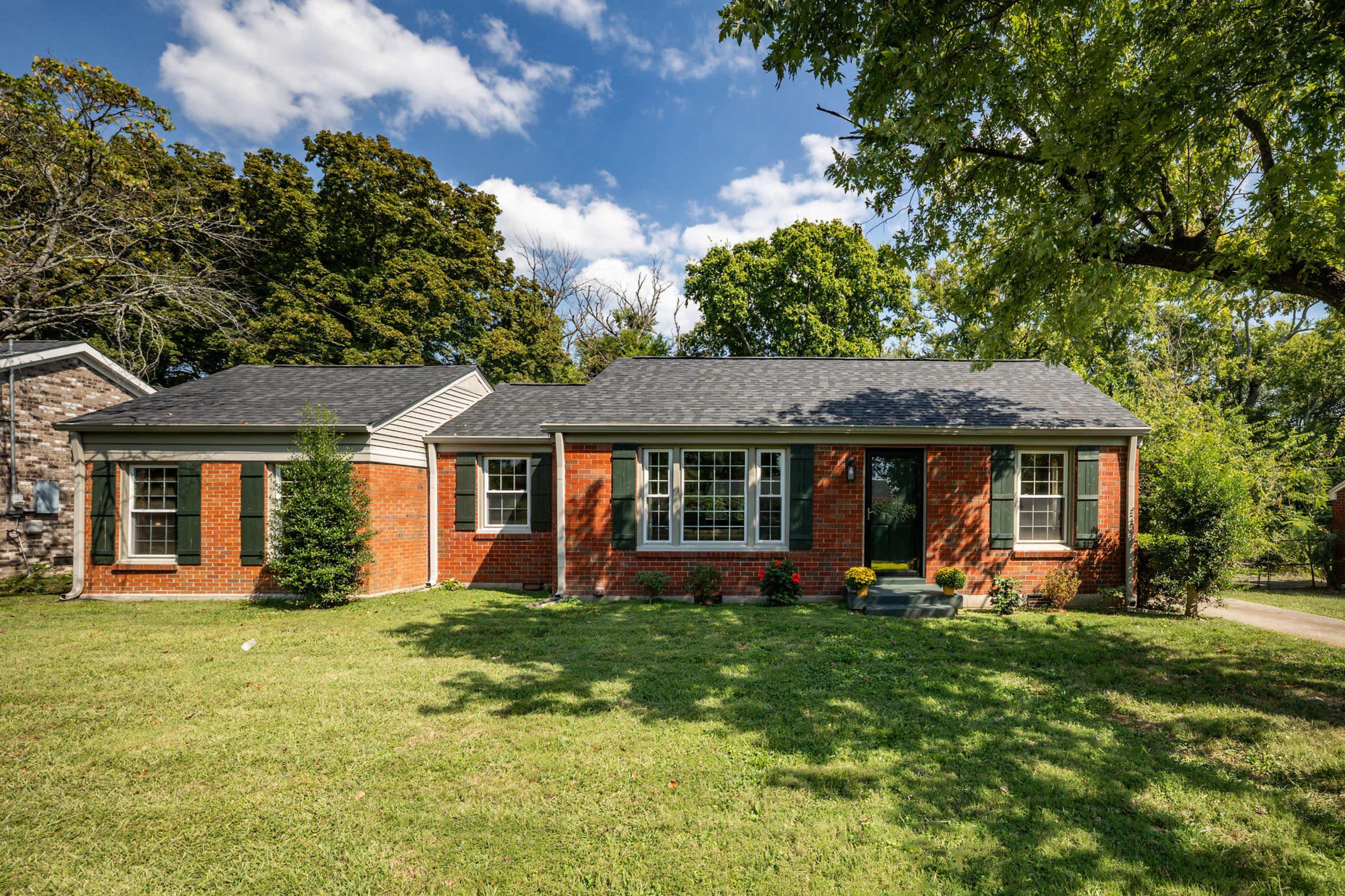 The image shows a red brick house with a dark roof, surrounded by a green lawn and trees, featuring multiple windows and a walkway.