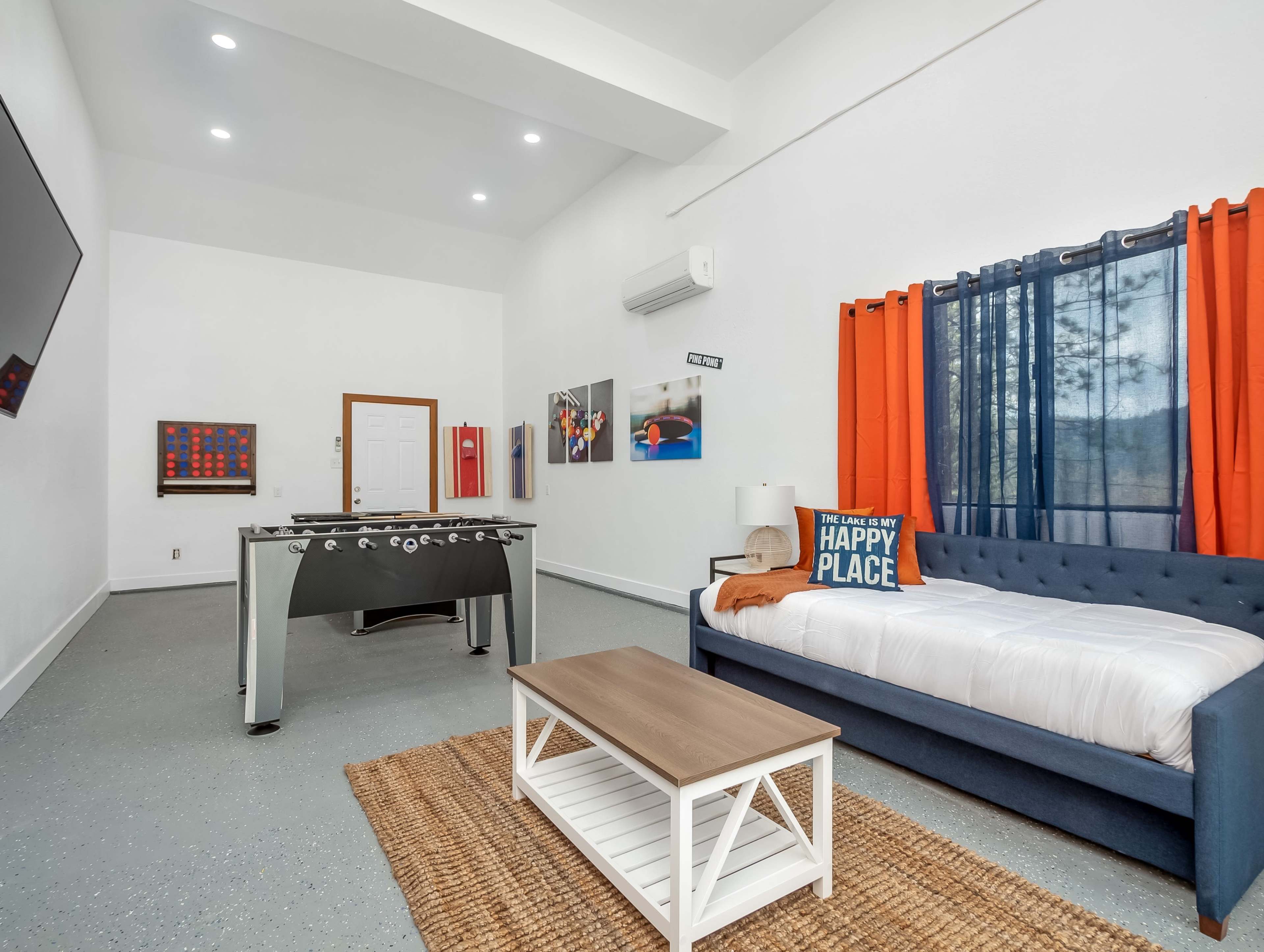 The room features a foosball table, a blue couch with a white blanket, a coffee table, and orange-accented curtains, all set against white walls and a gray floor.