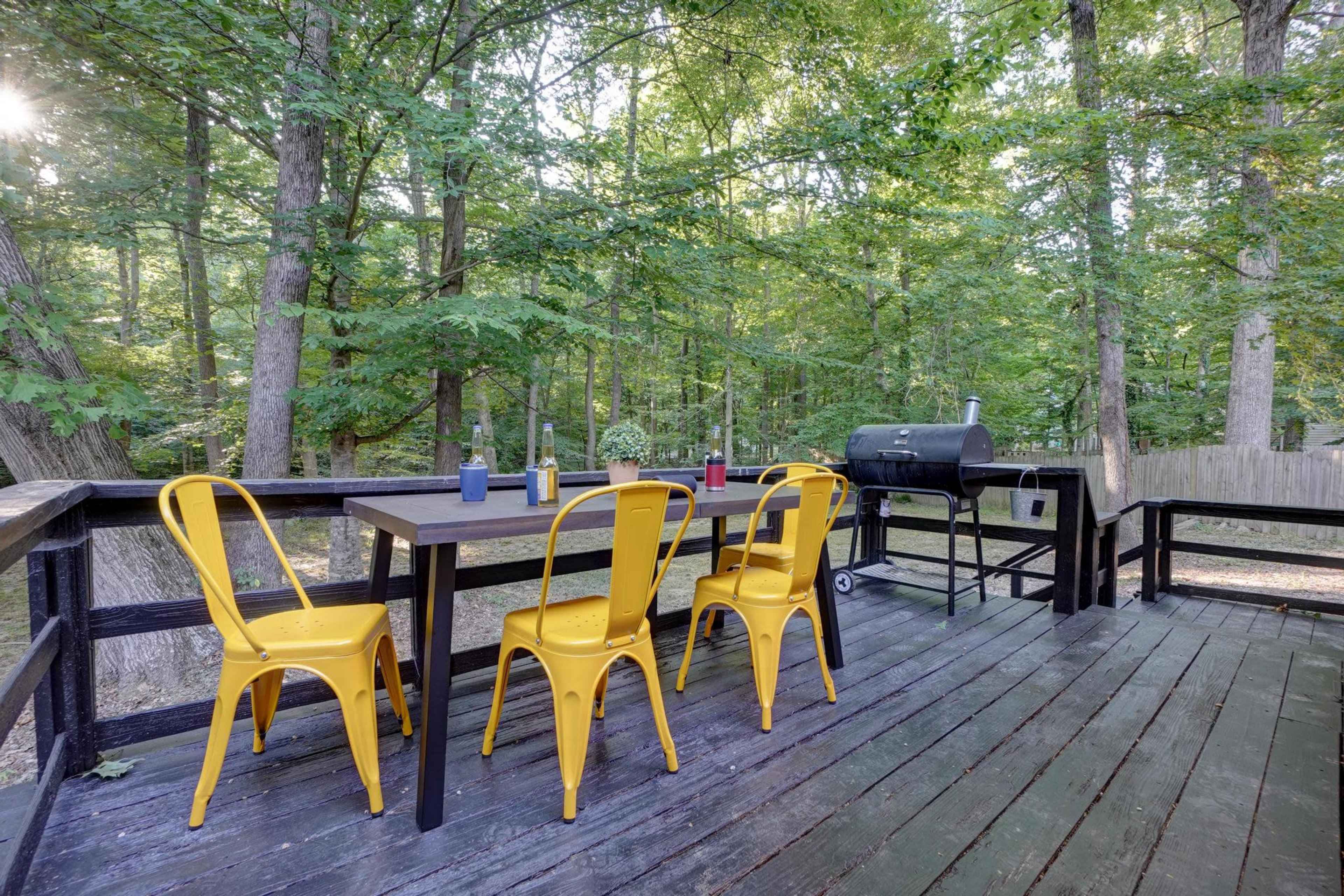 A wooden deck with a table and yellow chairs overlooks a wooded area, featuring a charcoal grill nearby.