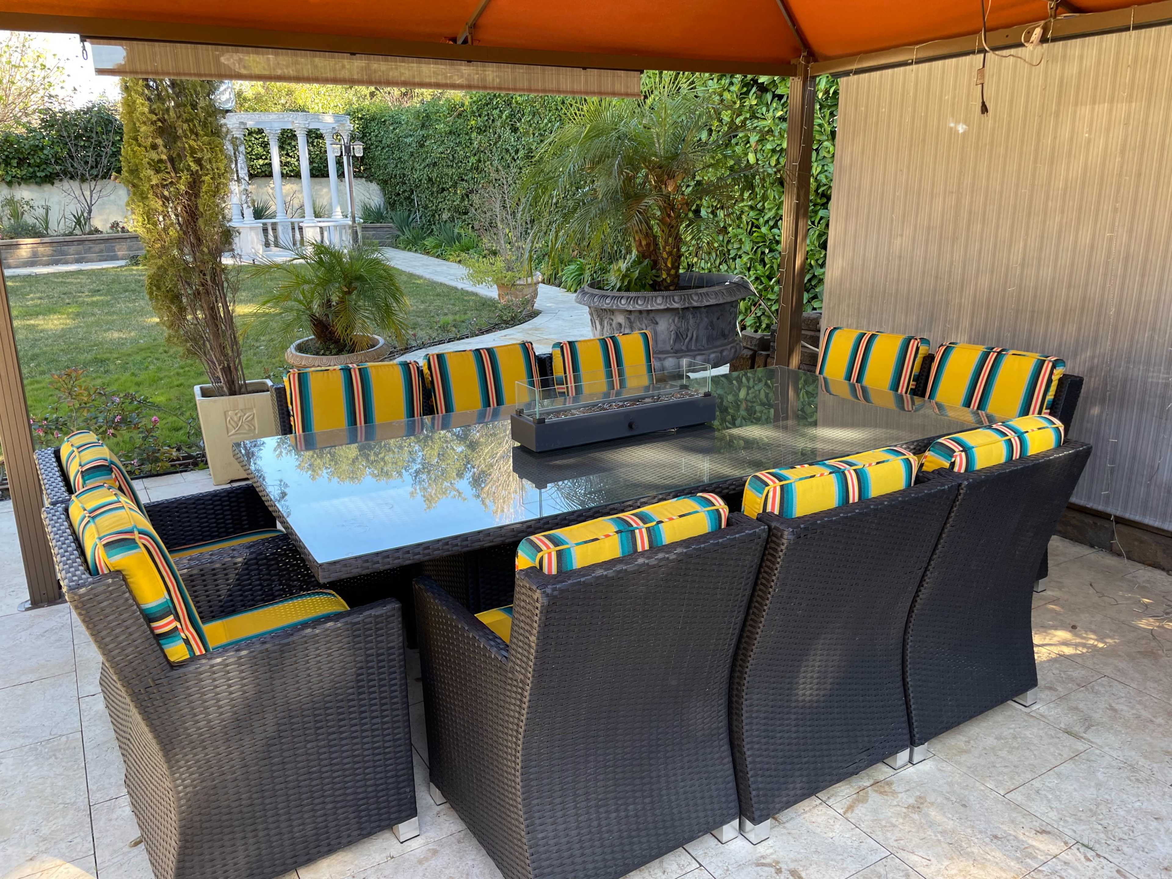 A large outdoor dining table with black wicker chairs and colorful striped cushions is set beneath a shaded gazebo in a garden.