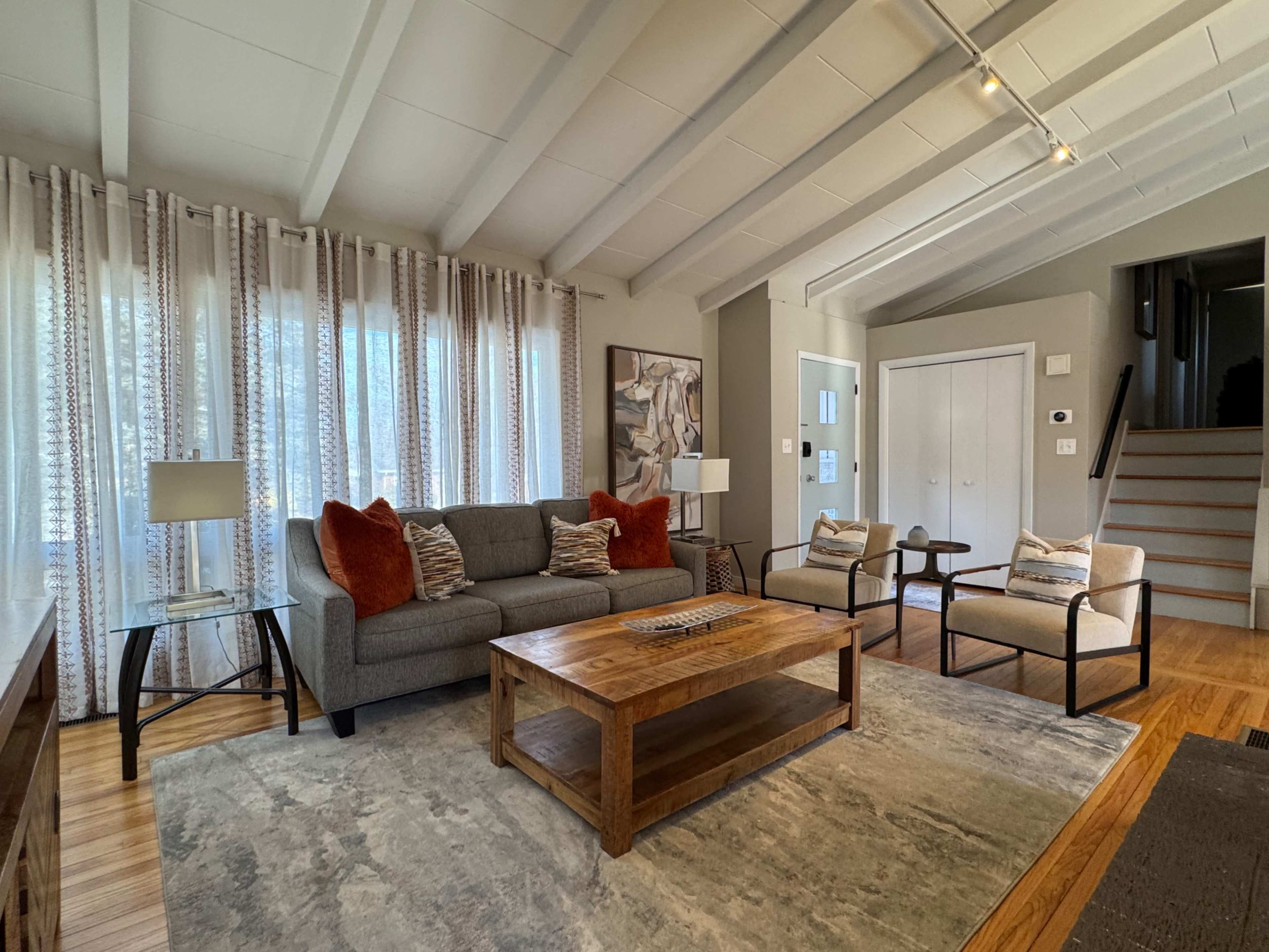 The living room features a gray sofa, wooden coffee table, and two armchairs, with large windows dressed in patterned curtains and exposed ceiling beams.